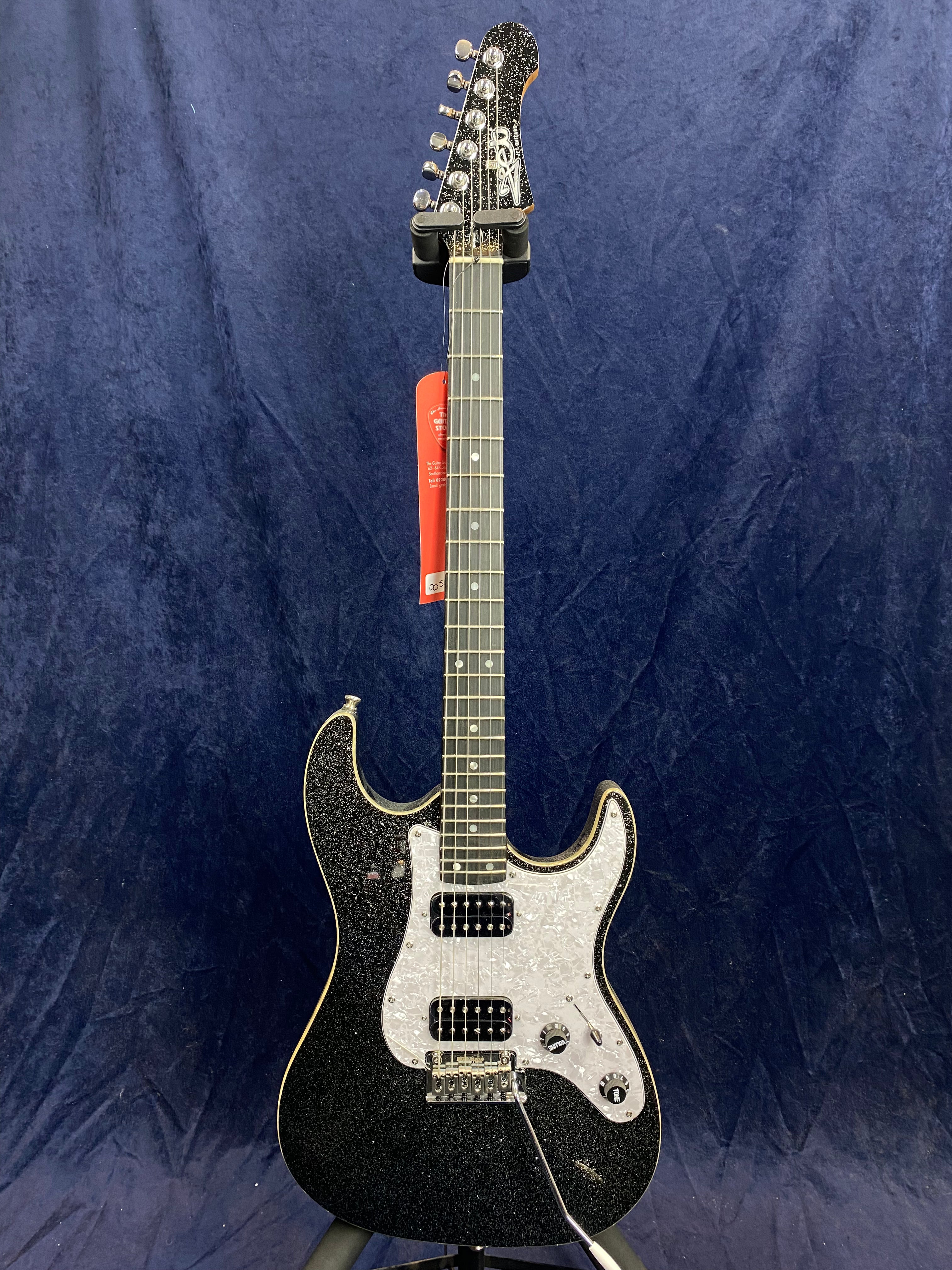 Black electric guitar with a sparkly finish, white pearloid pickguard, two pickups, a tremolo bridge, and a red tag on the neck, set against a dark background.