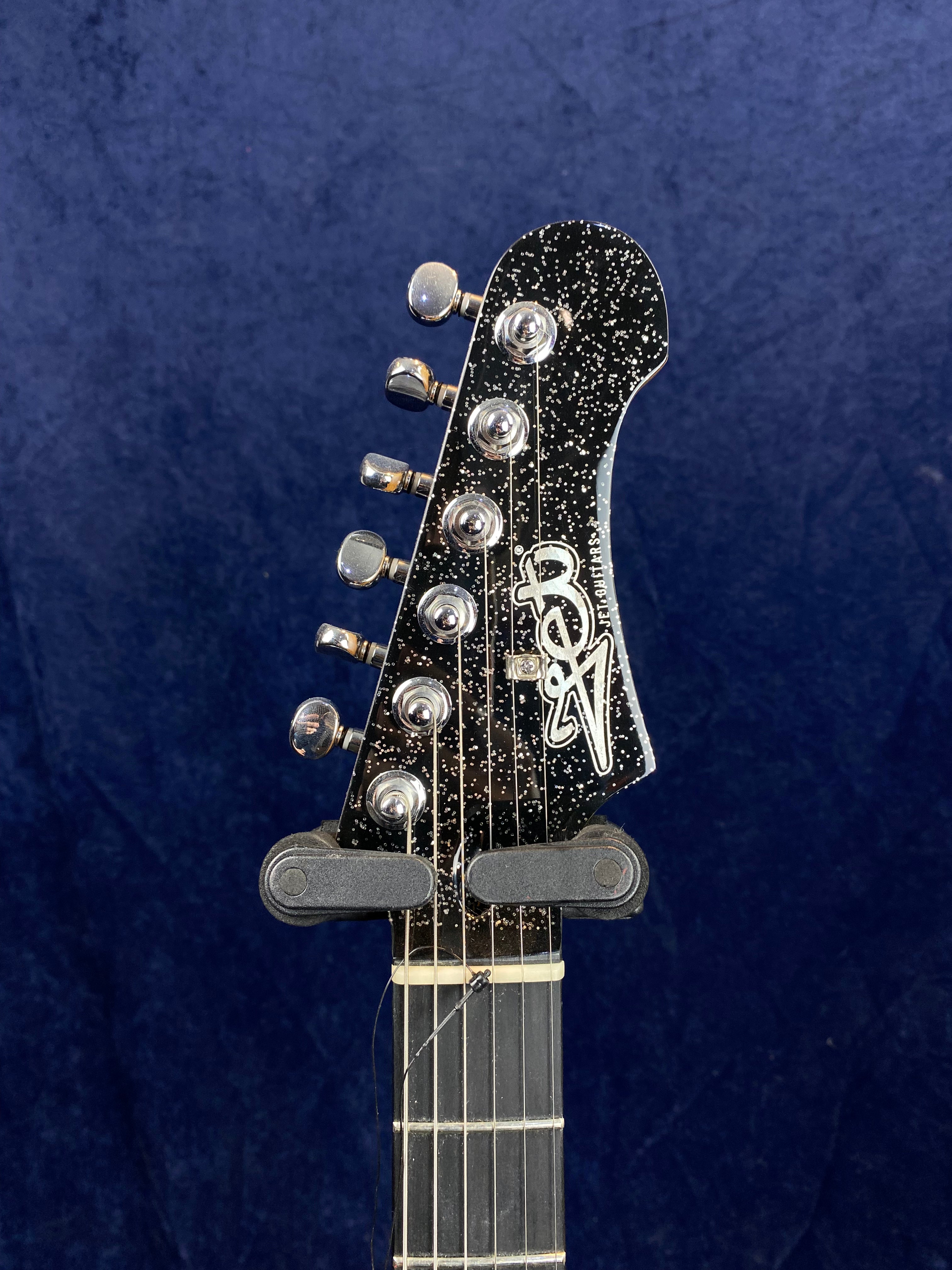 Electric guitar headstock with six chrome tuning pegs, black finish with white speckles, and a stylized white "BV" logo on the front.