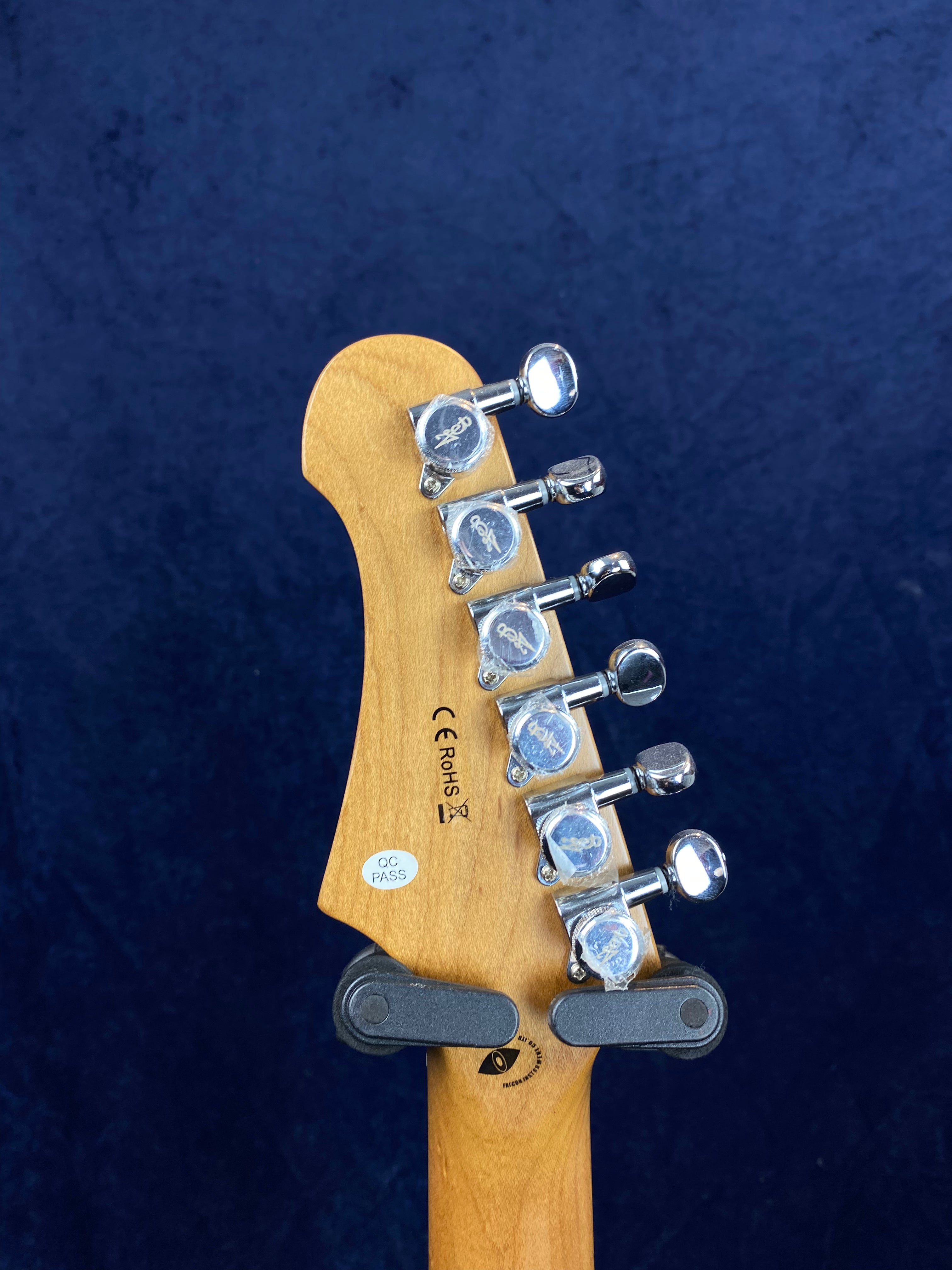 Six chrome tuning pegs line one side of a light wood electric guitar headstock, with certification and QC stickers visible on the back.