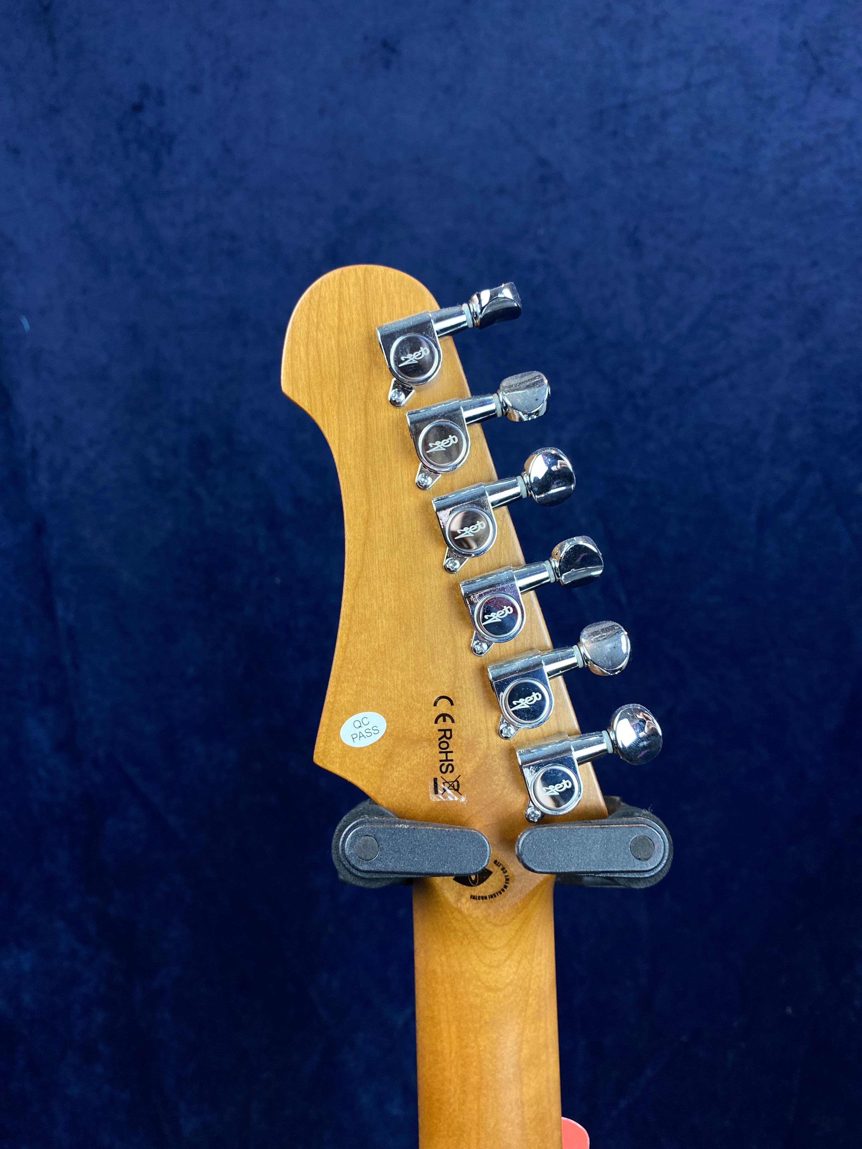 Closeup of electric guitar headstock, showing six chrome tuning pegs and certification stickers against a dark blue background.