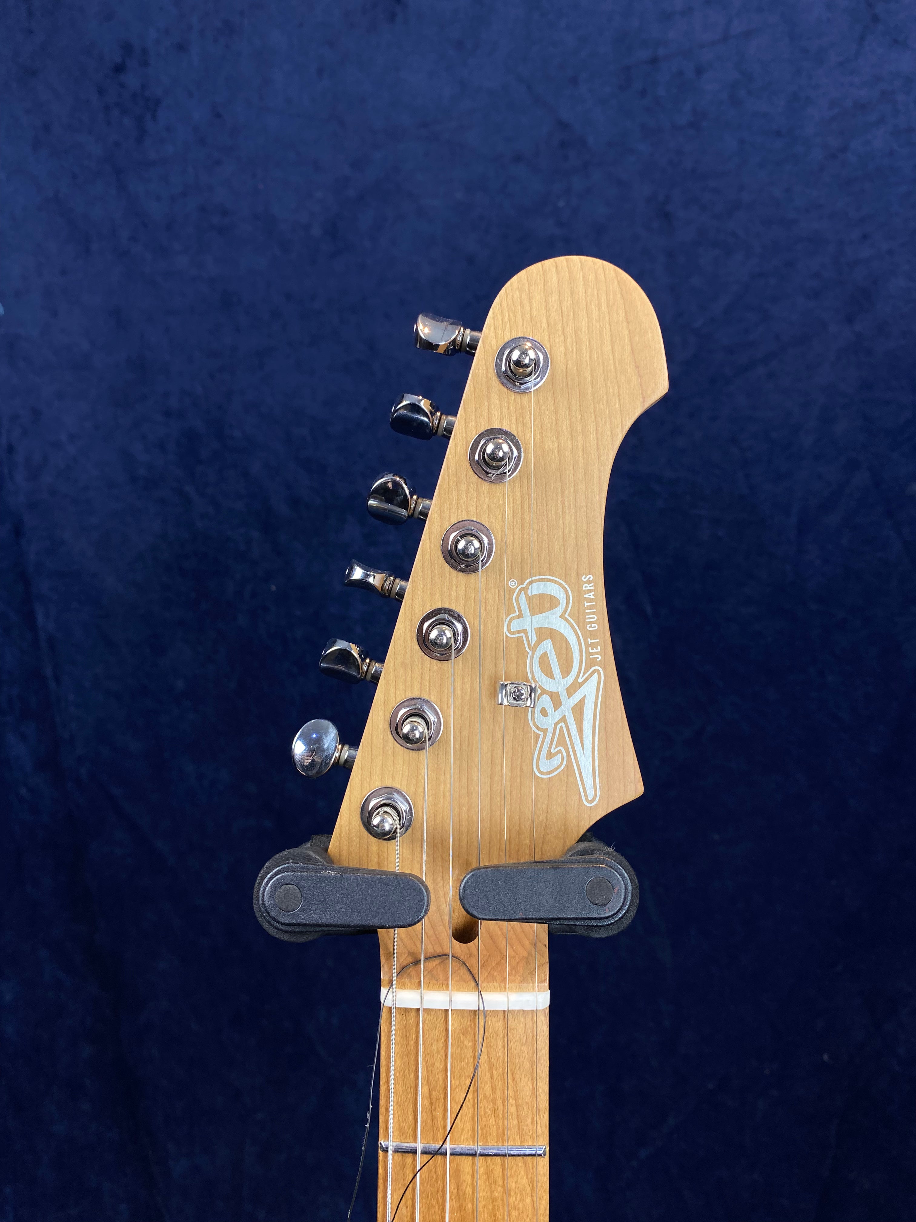 Headstock of a Jet brand electric guitar with six chrome tuning pegs and visible strings against a dark blue background.