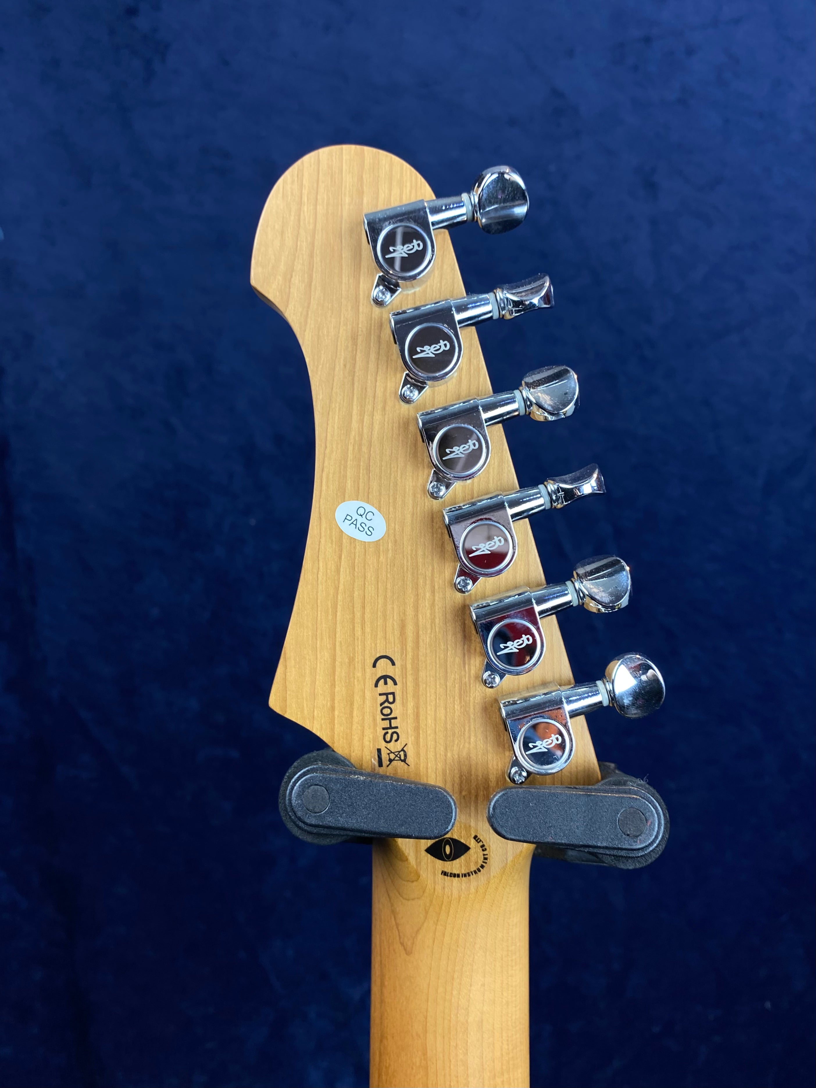 Six chrome tuning pegs on a light wood electric guitar headstock, with a QC Pass sticker and compliance markings visible, set against a dark blue background.