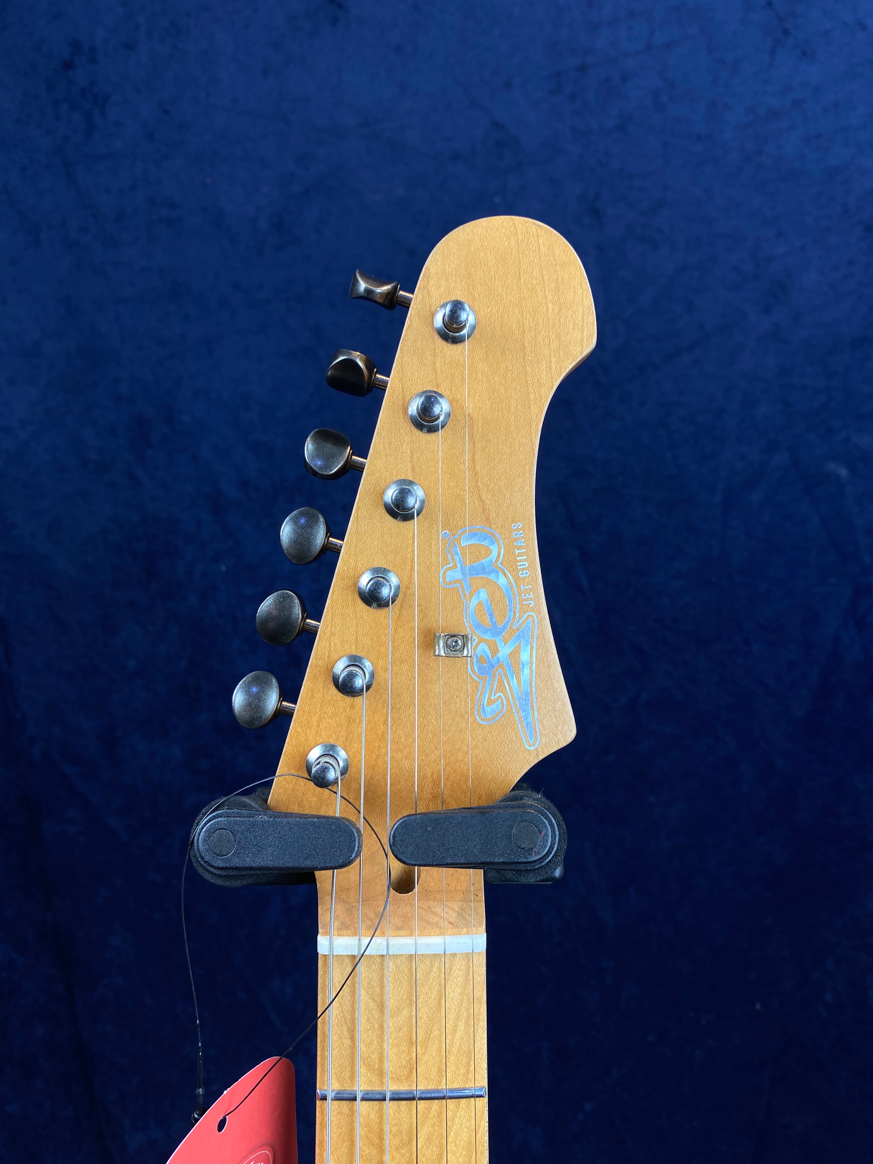 Natural wood electric guitar headstock with six metal tuning pegs facing left, a blue logo, and a black hanging holder against a dark blue background.
