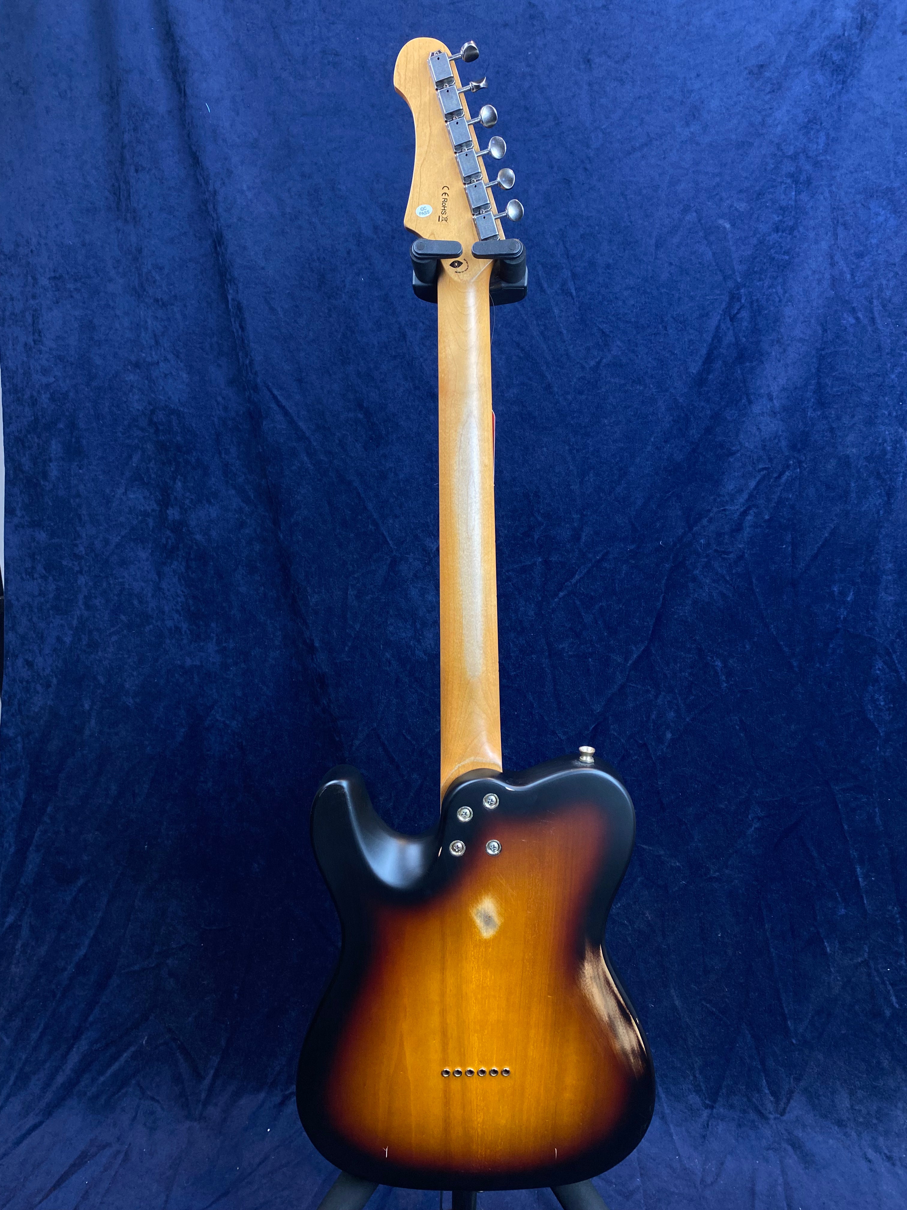 Sunburst-finish electric guitar seen from the back, showing tuning pegs, natural wood neck, rounded body, and metal screws on a dark blue background.