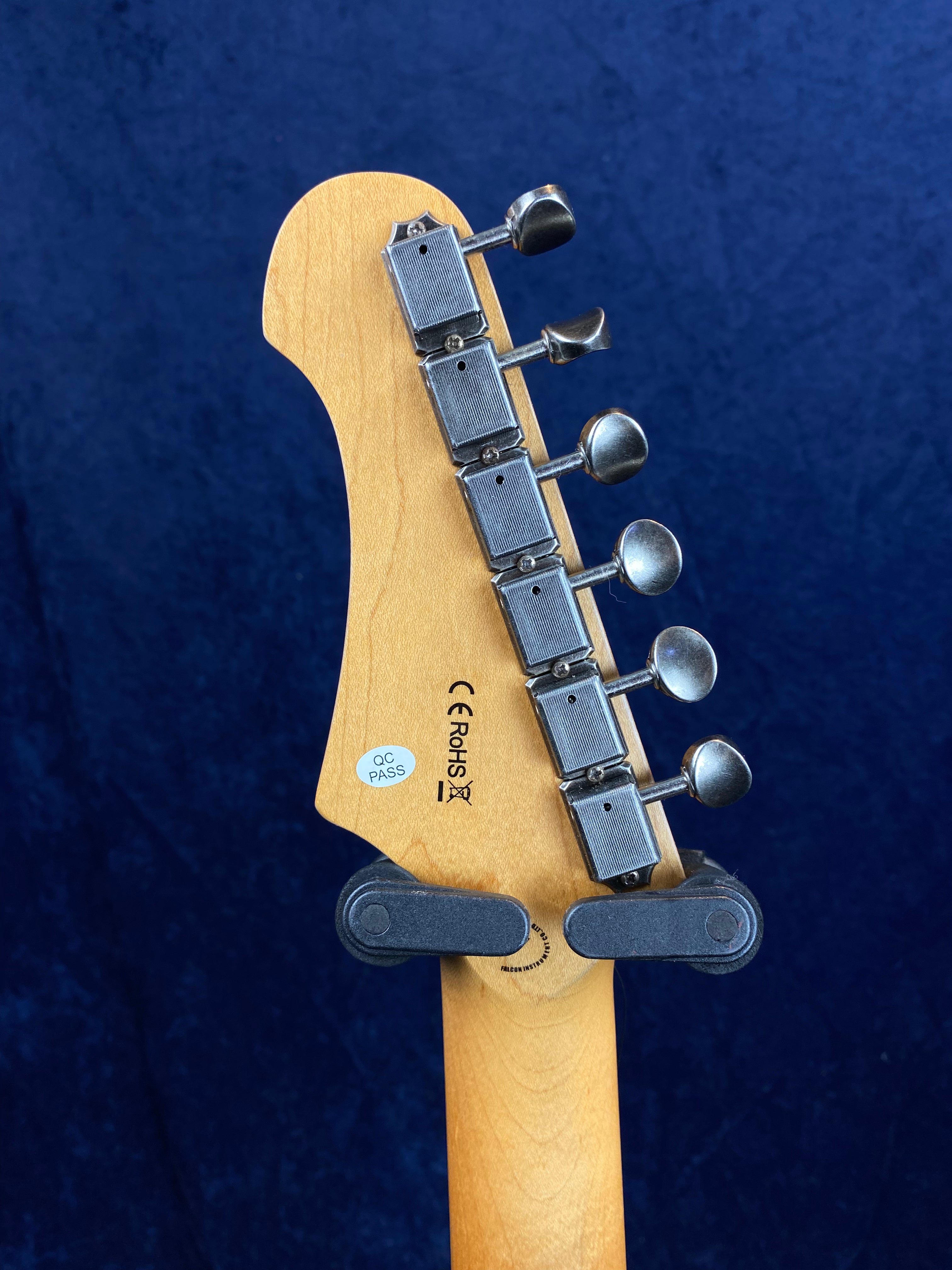 Six metal tuning pegs line one side of a light wood electric guitar headstock, with a QC PASS sticker and CE RoHS markings visible.