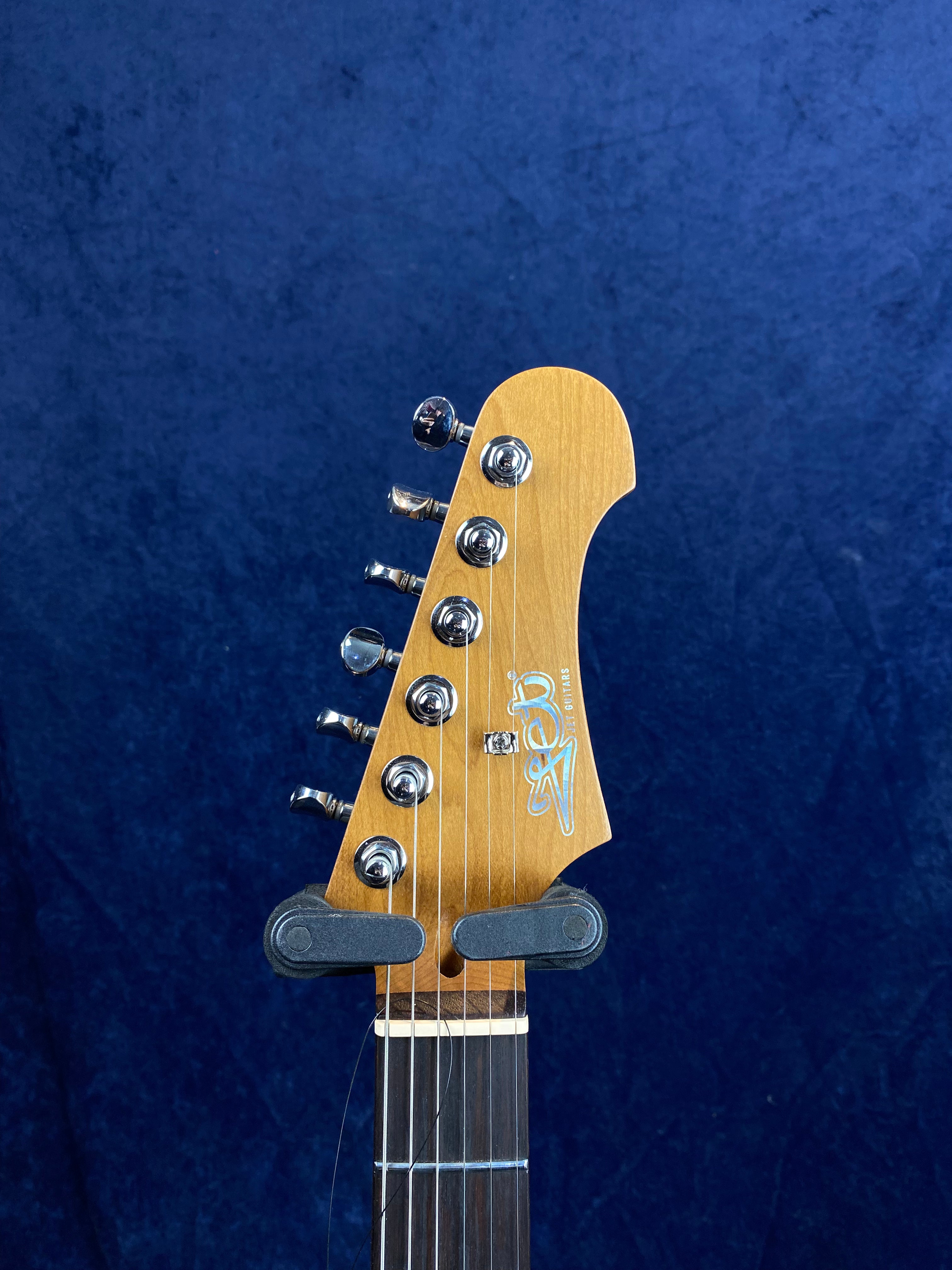 Electric guitar headstock with six silver tuning pegs, maple finish, and "SEW Guitars" logo, set against a dark blue background.