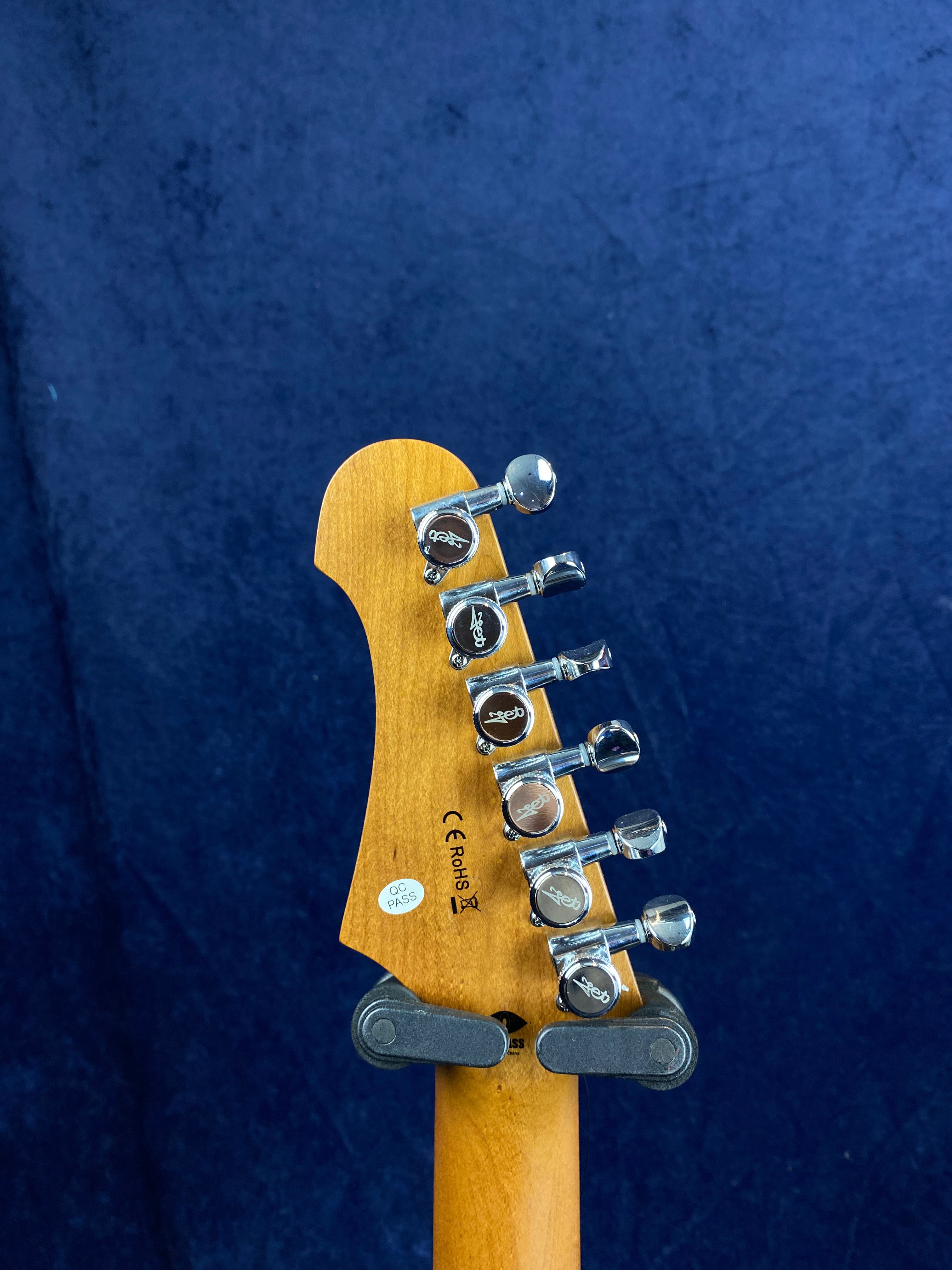 Six chrome tuning pegs line the back of a wooden electric guitar headstock, with certification stickers and a blue background.