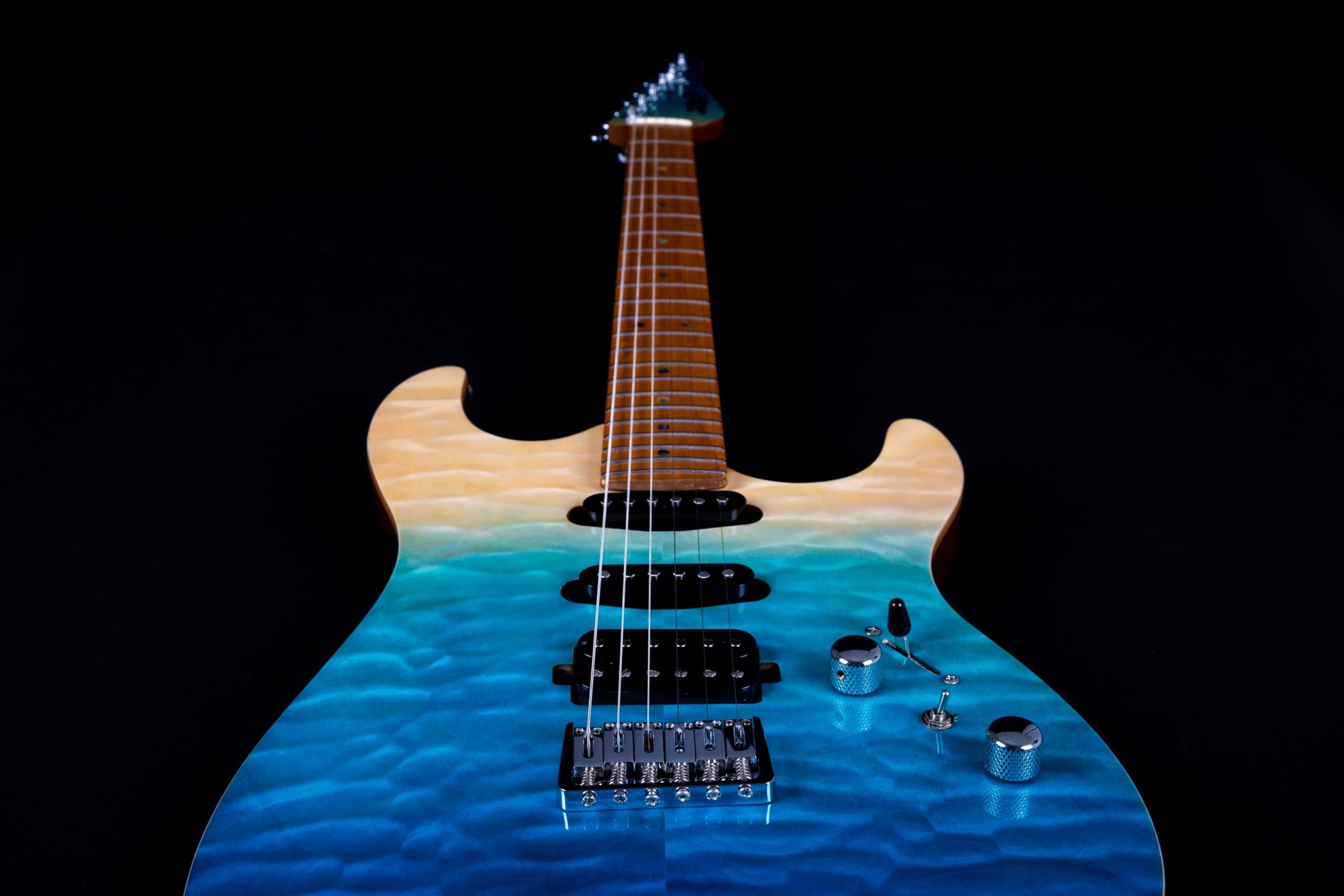Electric guitar with a quilted finish, fading from sandy tan at the top to blue at the bottom, chrome controls, three pickups, and a maple neck.