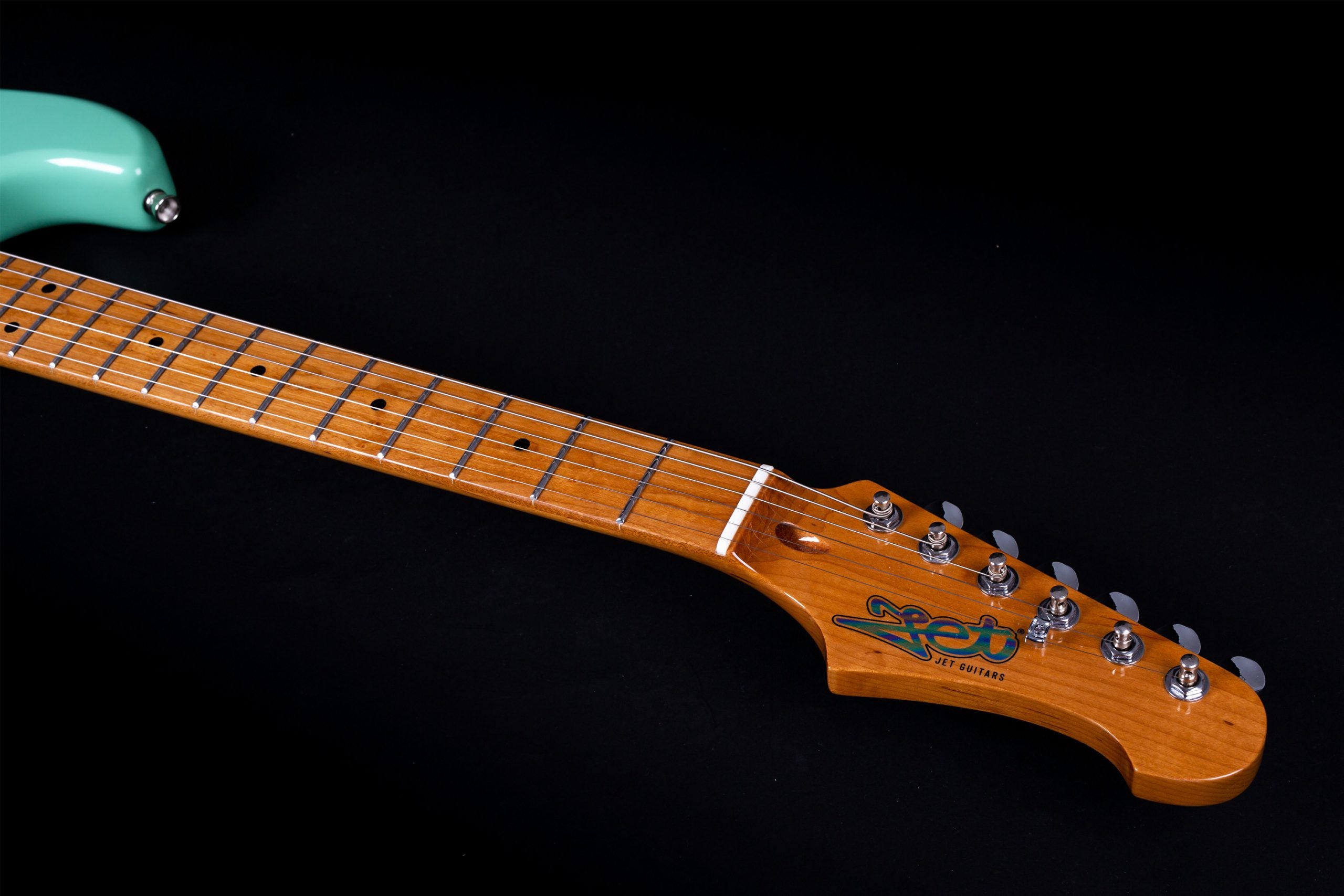 A maple neck and headstock of a Jet brand electric guitar with six tuning pegs, set against a black background. The body is seafoam green.