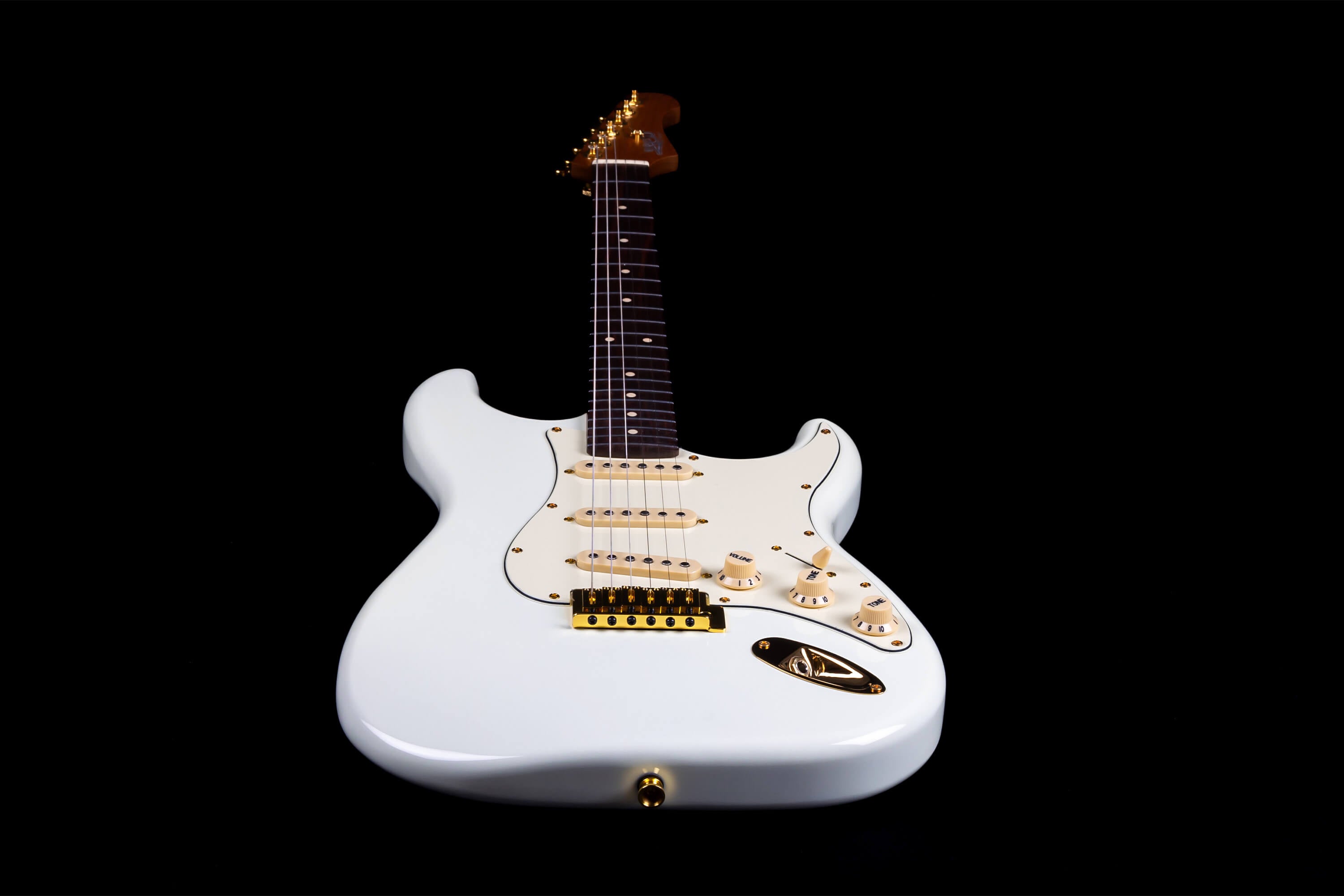White electric guitar with gold hardware, cream pickguard, three single-coil pickups, and a dark fretboard, viewed from the body end against a black background.