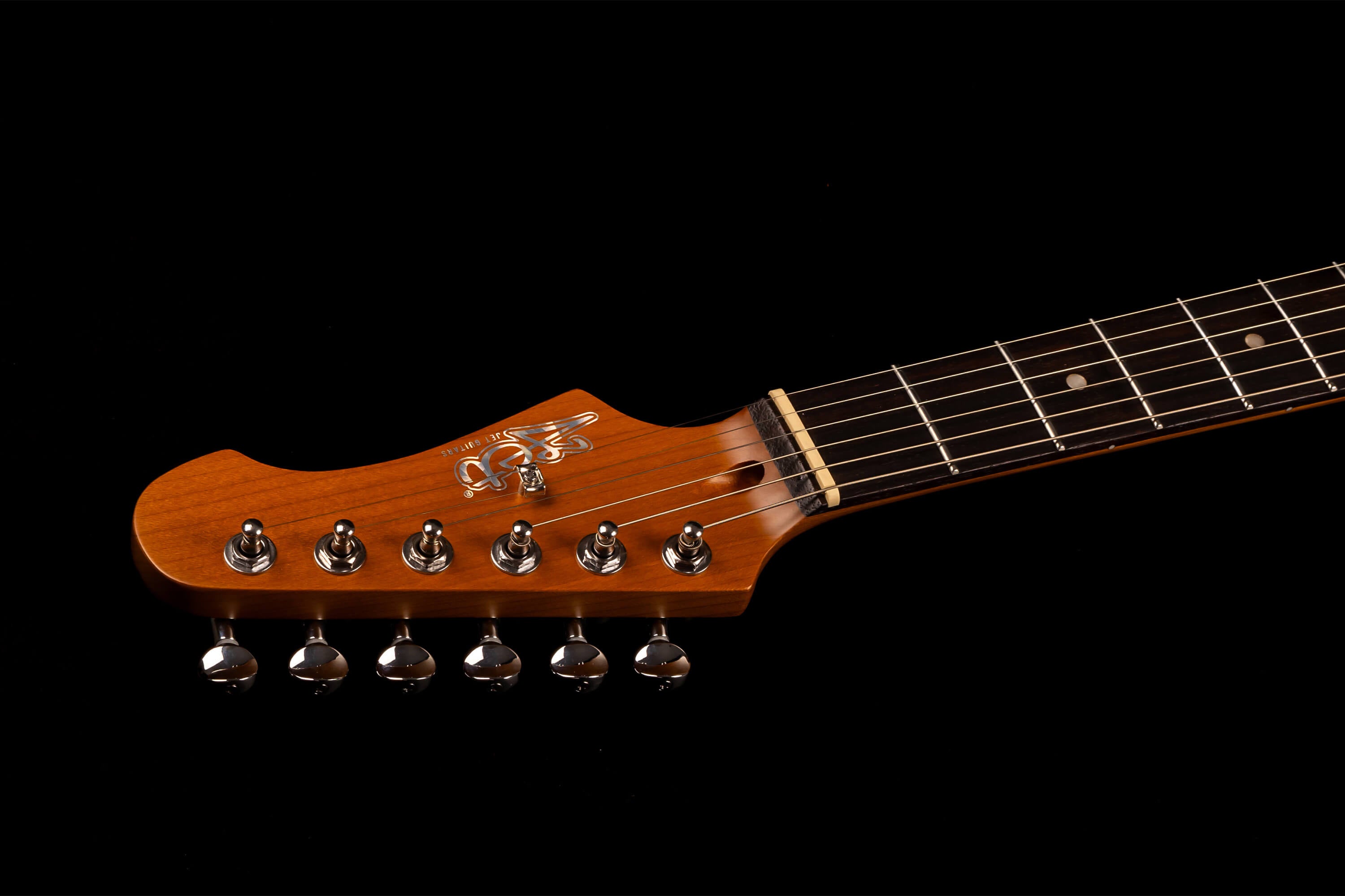 Light brown electric guitar headstock with six tuning pegs, metallic logo, and part of a dark fingerboard with metal frets and white dot inlays.