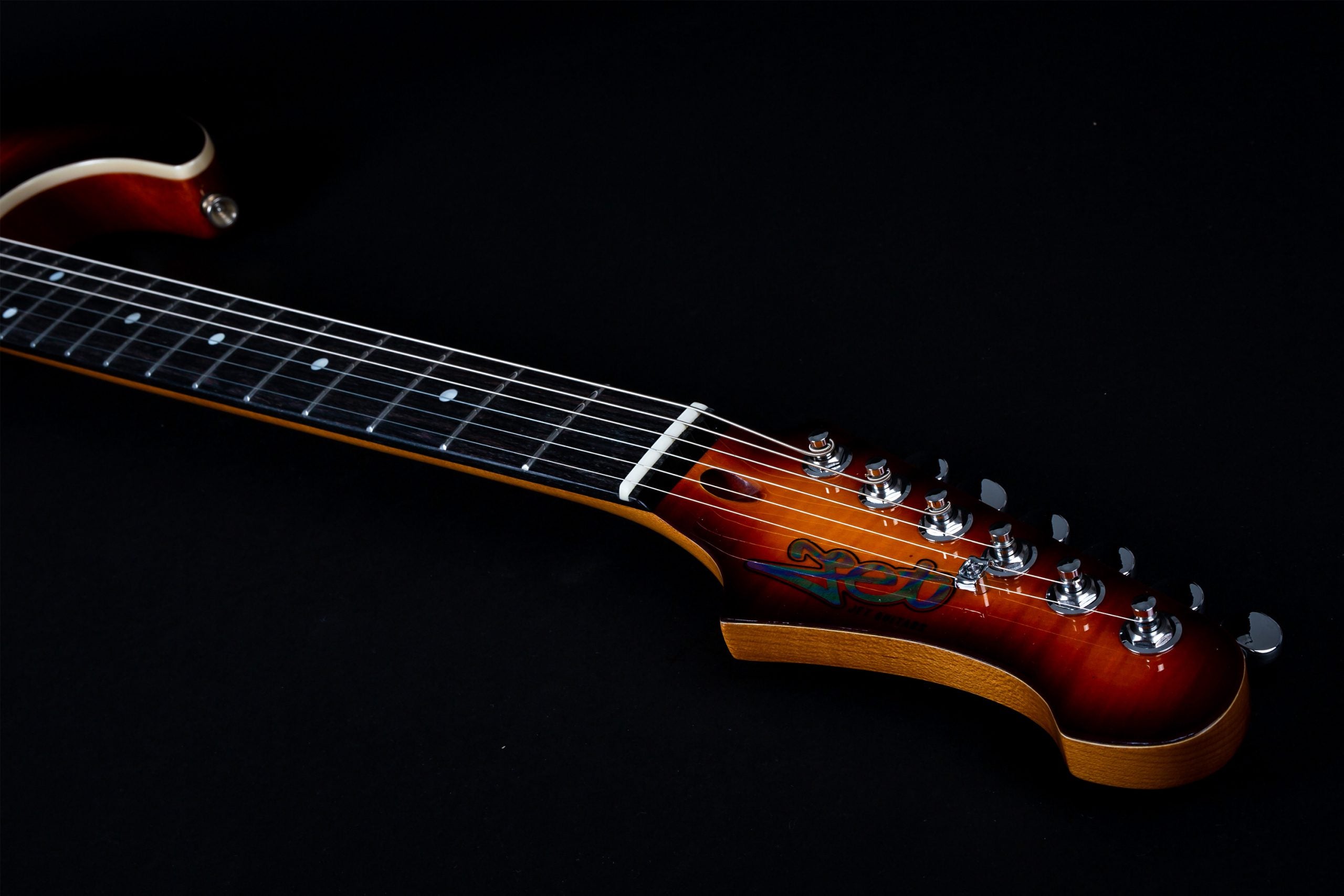 Sunburst electric guitar neck and headstock with six tuning pegs, white fretboard inlays, and a colorful logo on the head, set against black.
