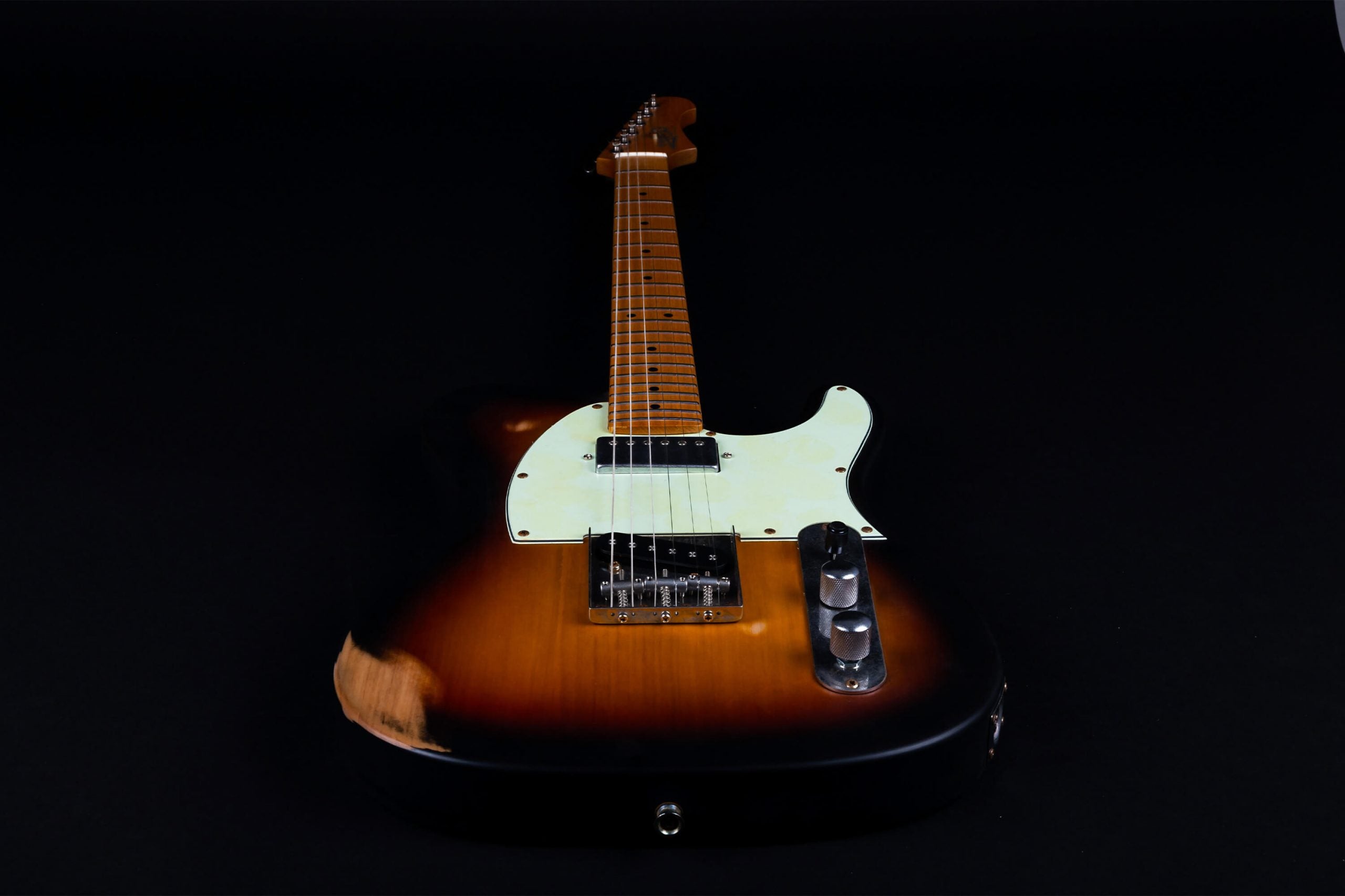 Sunburst-finish electric guitar with a white pickguard, maple neck, metal bridge, two control knobs, and visible wear on the lower left body.
