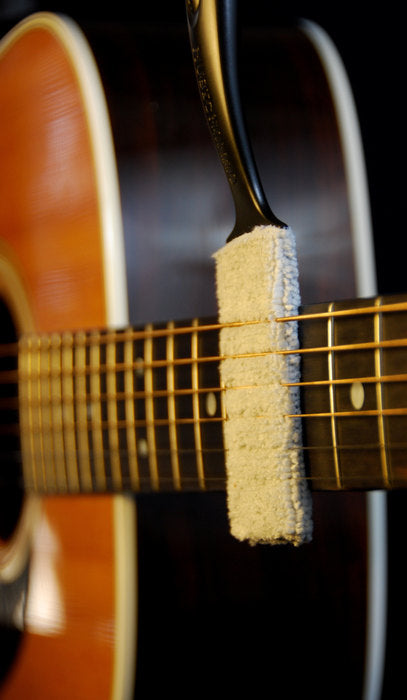 A white cleaning tool with a handle is pressed against the strings and fretboard of an acoustic guitar, likely for removing dust or grime.