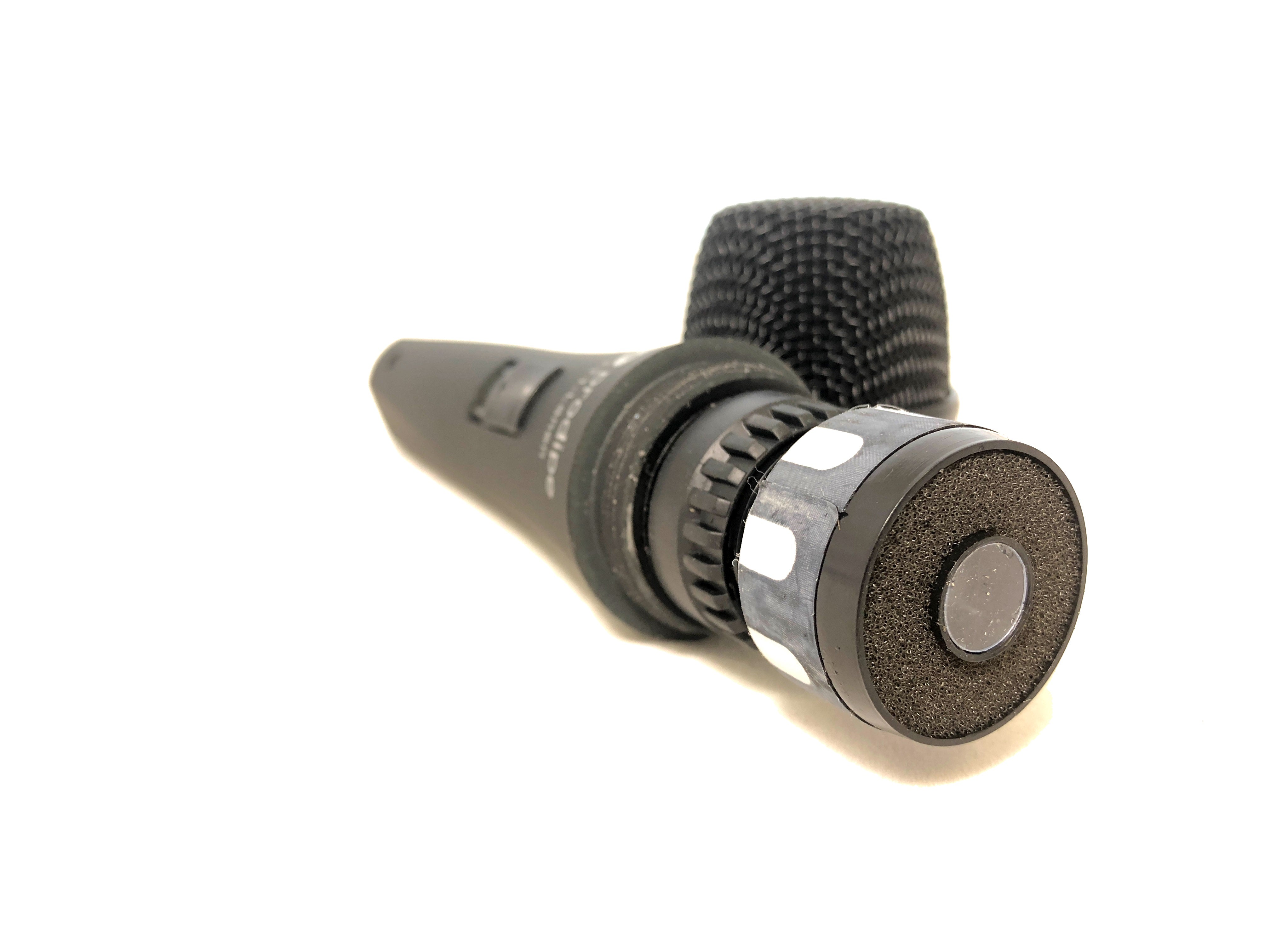 Black handheld microphone with a mesh head, an on-off switch on the body, and a circular foam-covered end resting on a white surface.