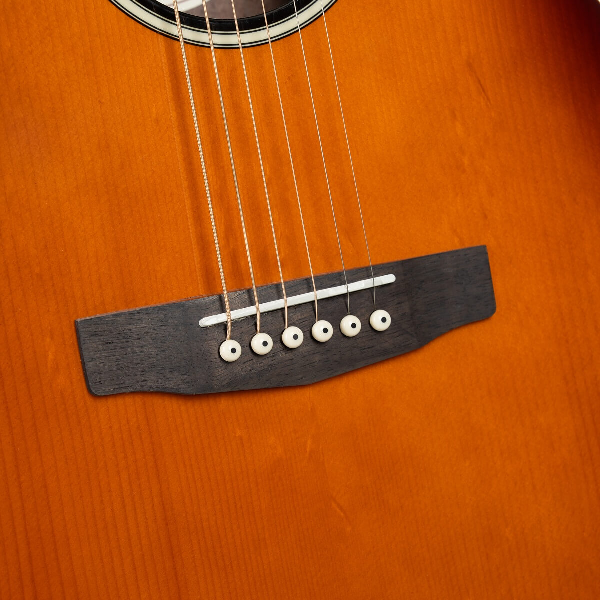 Close-up of an acoustic guitar body showing six steel strings, dark wood bridge, and white bridge pins on a glossy orange-brown finish.