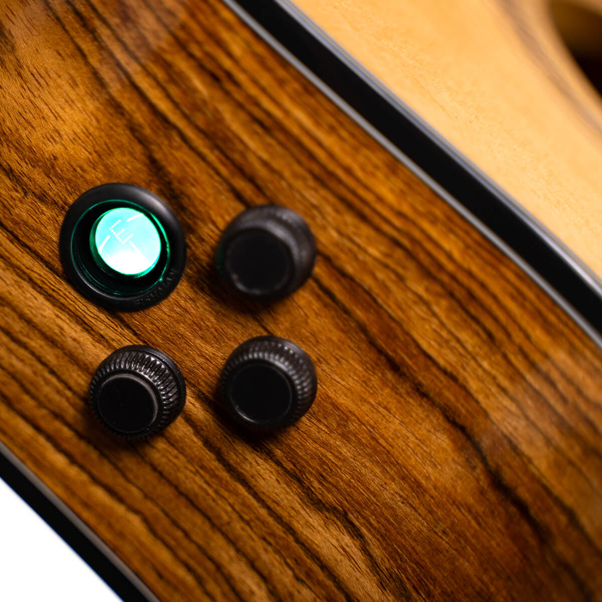 Close-up of an acoustic guitar's side panel showing a green-lit electronic tuner button and three black control knobs on a wood grain finish.