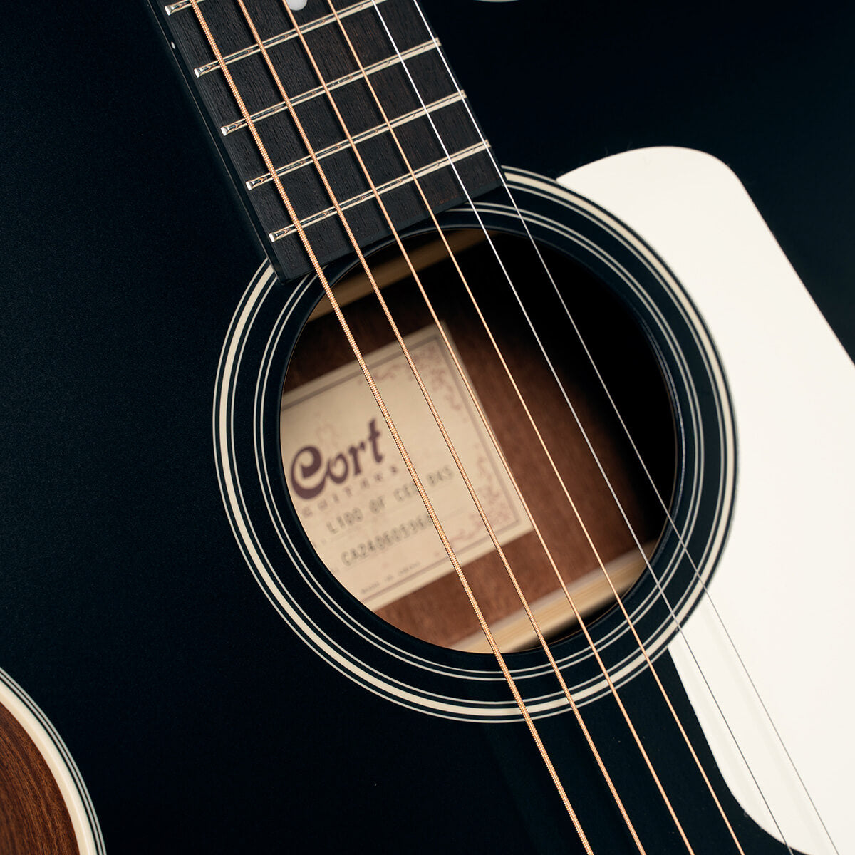 Close-up of a Cort acoustic guitar soundhole, showing steel strings, part of the fretboard, and a white pickguard.