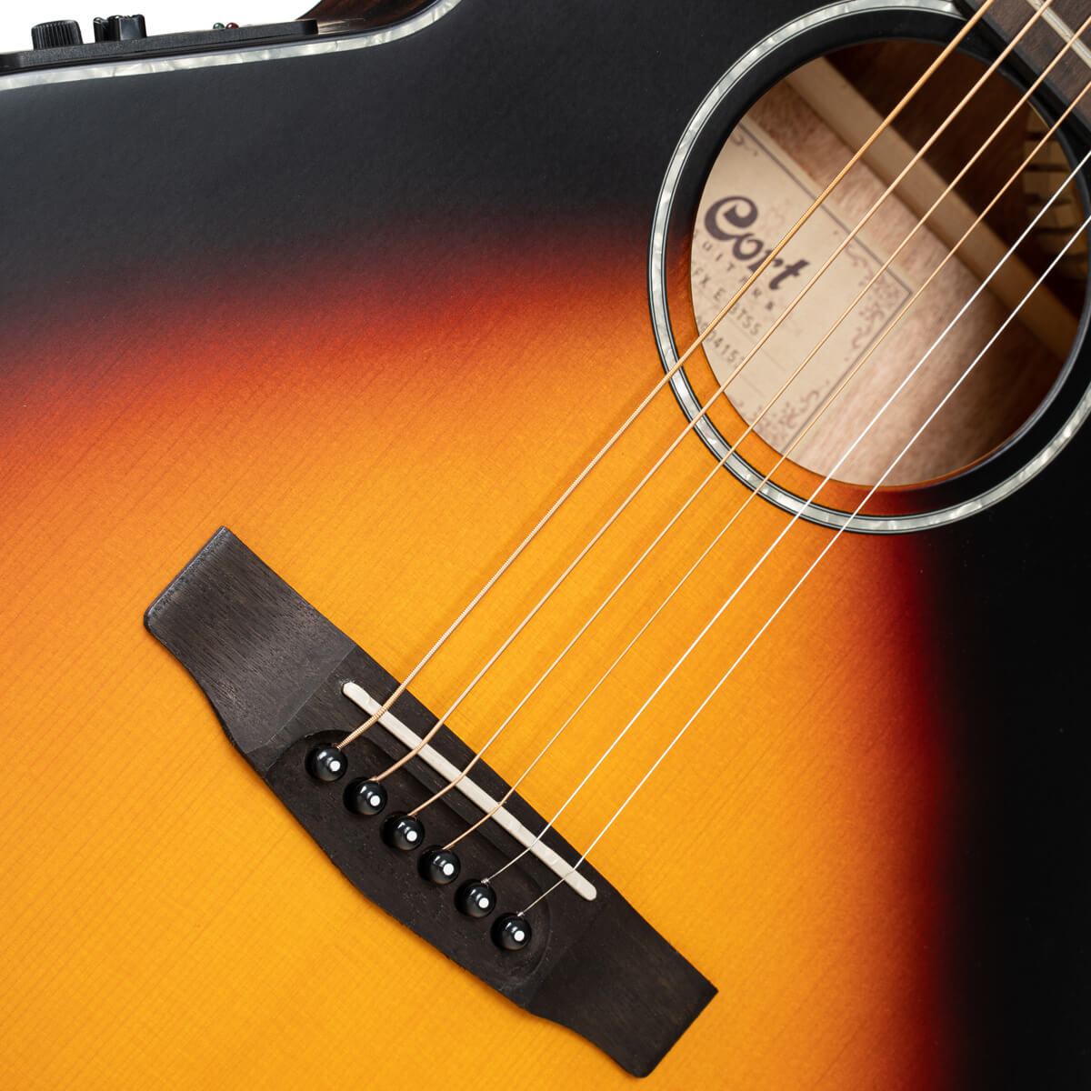 Sunburst finish acoustic guitar close-up showing six strings, black bridge with pins, and round sound hole with the Cort brand label inside.
