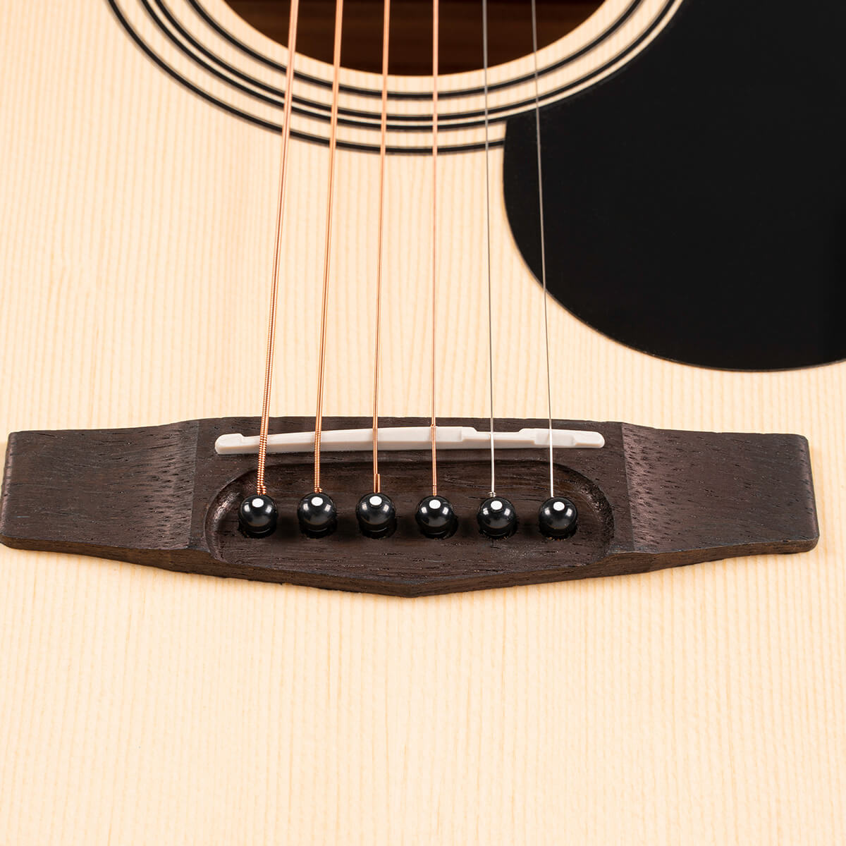 Close-up of an acoustic guitar bridge with six strings and black bridge pins on a light wood body. Part of the sound hole and pickguard are visible.