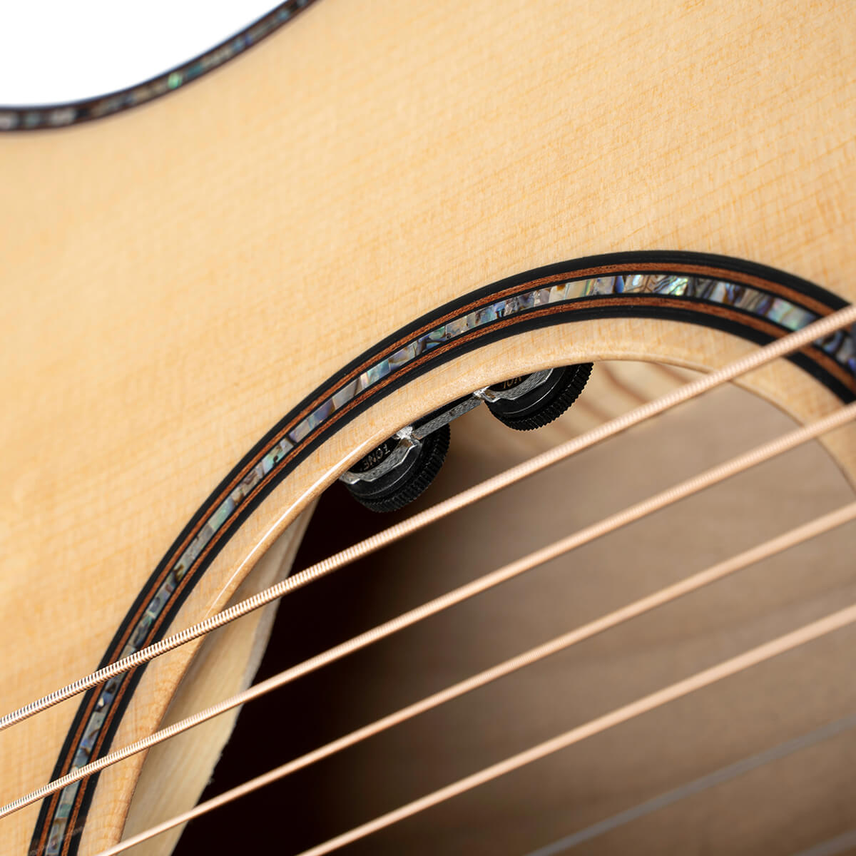 Close-up of an acoustic guitar’s soundhole with abalone inlay, showing steel strings and two black control knobs just inside the hole.
