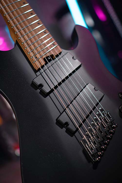 Close-up of a matte black electric guitar with eight strings, two rectangular pickups, and a multi-saddle bridge, viewed at an angle.