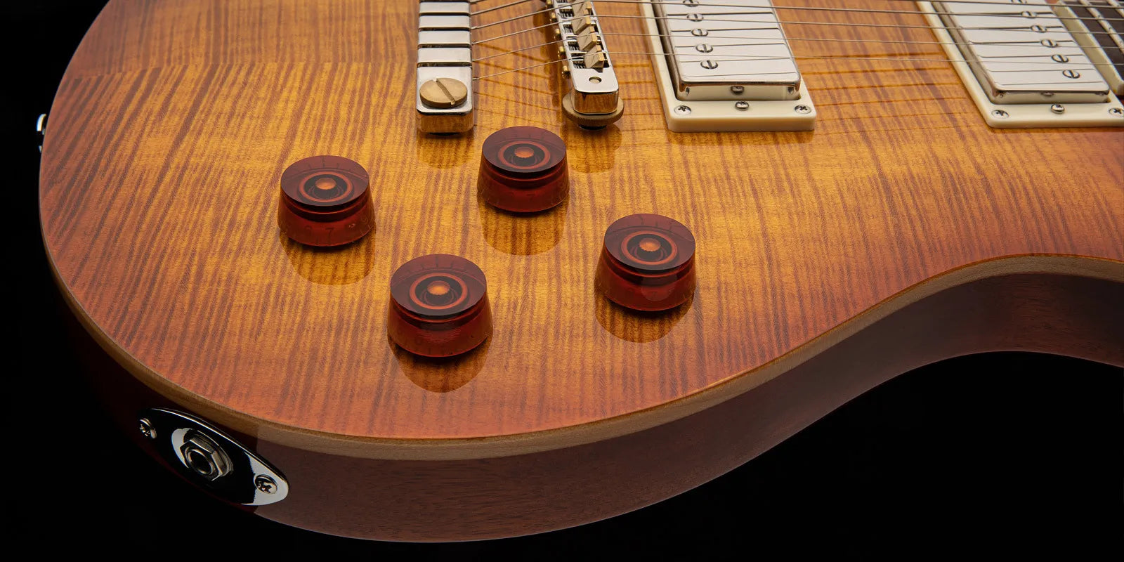 Sunburst finish electric guitar close-up, showing four amber control knobs, metal input jack, and part of the bridge with string detail.