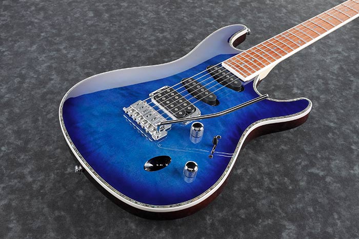 Electric guitar with a glossy blue body, three pickups, chrome knobs, a whammy bar, and a light-colored fingerboard on a dark marbled surface.