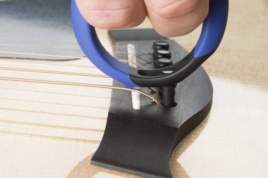 A hand uses a blue and black tool to lift a bridge pin from an acoustic guitar, helping with cleaning or string maintenance.