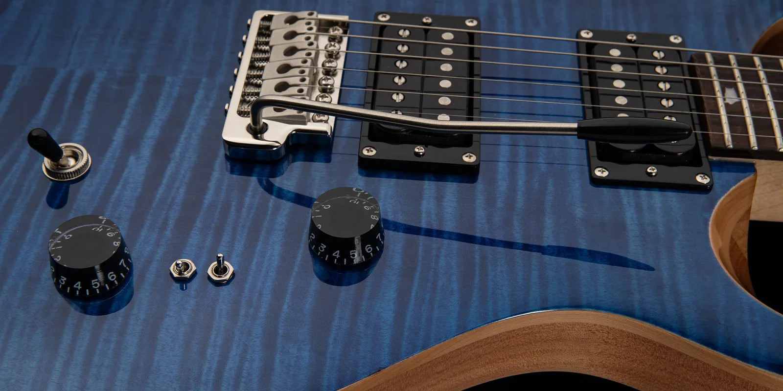 Blue electric guitar body with black knobs, toggle switches, two humbucker pickups, a tremolo bridge, and a whammy bar; wood edge visible.