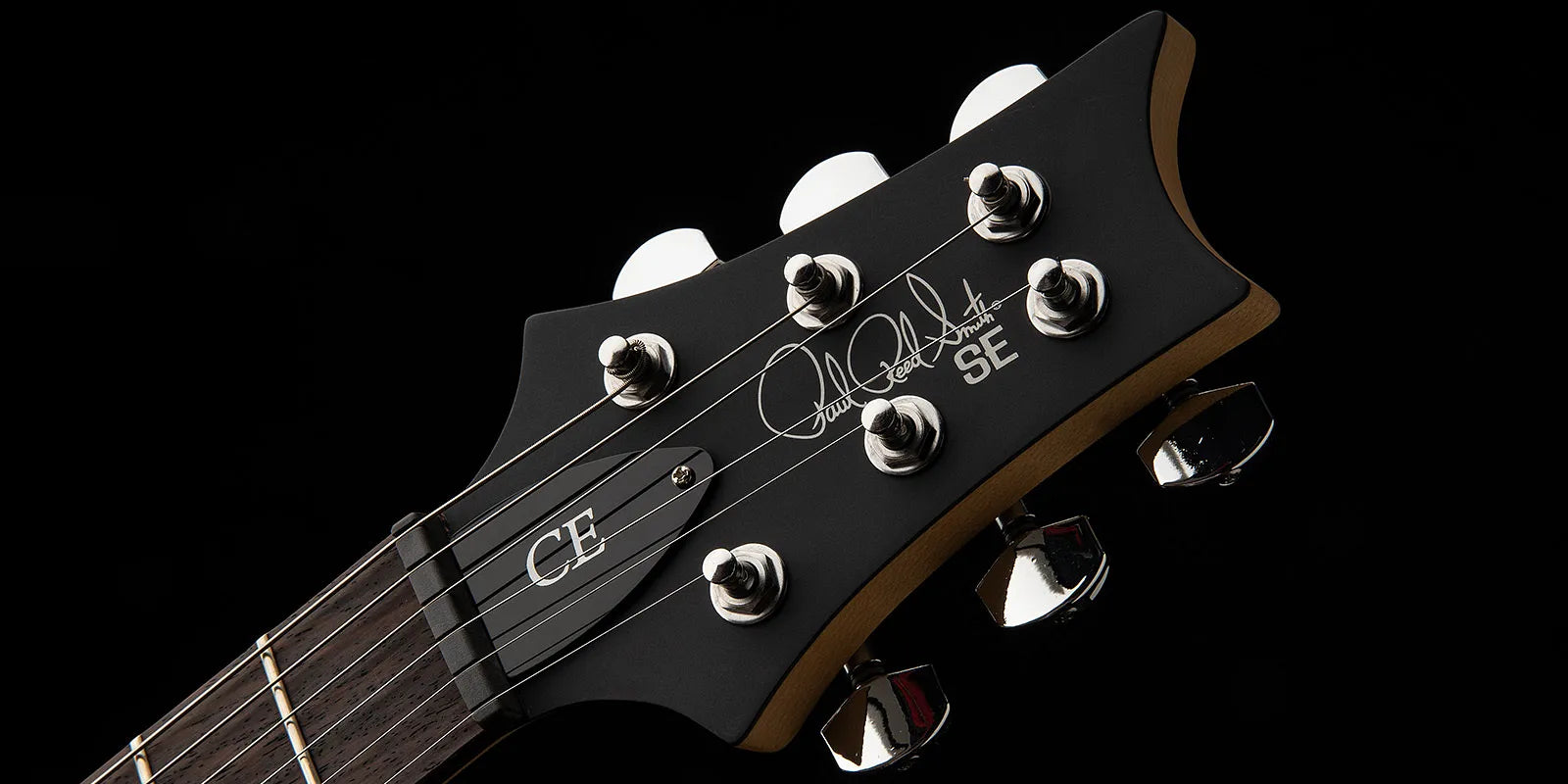 Electric guitar headstock with six chrome tuning pegs, “CE” truss rod cover, and “Paul Reed Smith SE” signature on matte black finish.