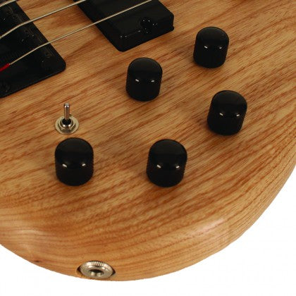Natural wood bass guitar body showing five black control knobs, a small toggle switch, and a metal input jack near the edge.