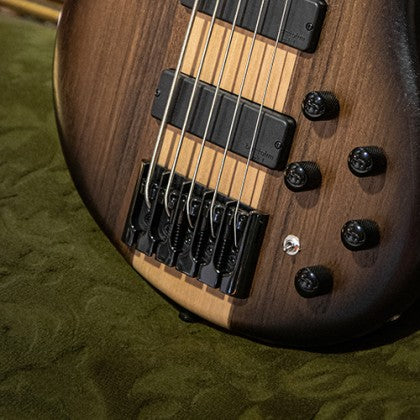 Five-string bass guitar body in natural wood finish with black hardware, bridge, pickups, and five control knobs on a textured green surface.