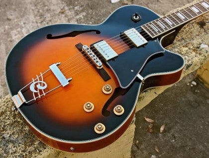 Sunburst finish electric guitar with f-holes, four control knobs, chrome bridge, and black pickguard resting on a stone surface.