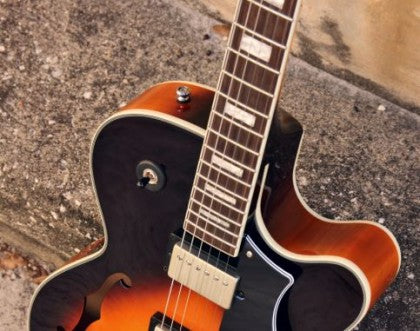 Sunburst semi-hollow electric guitar with f-hole, cream binding, metal controls, and a wooden neck rests on concrete pavement.