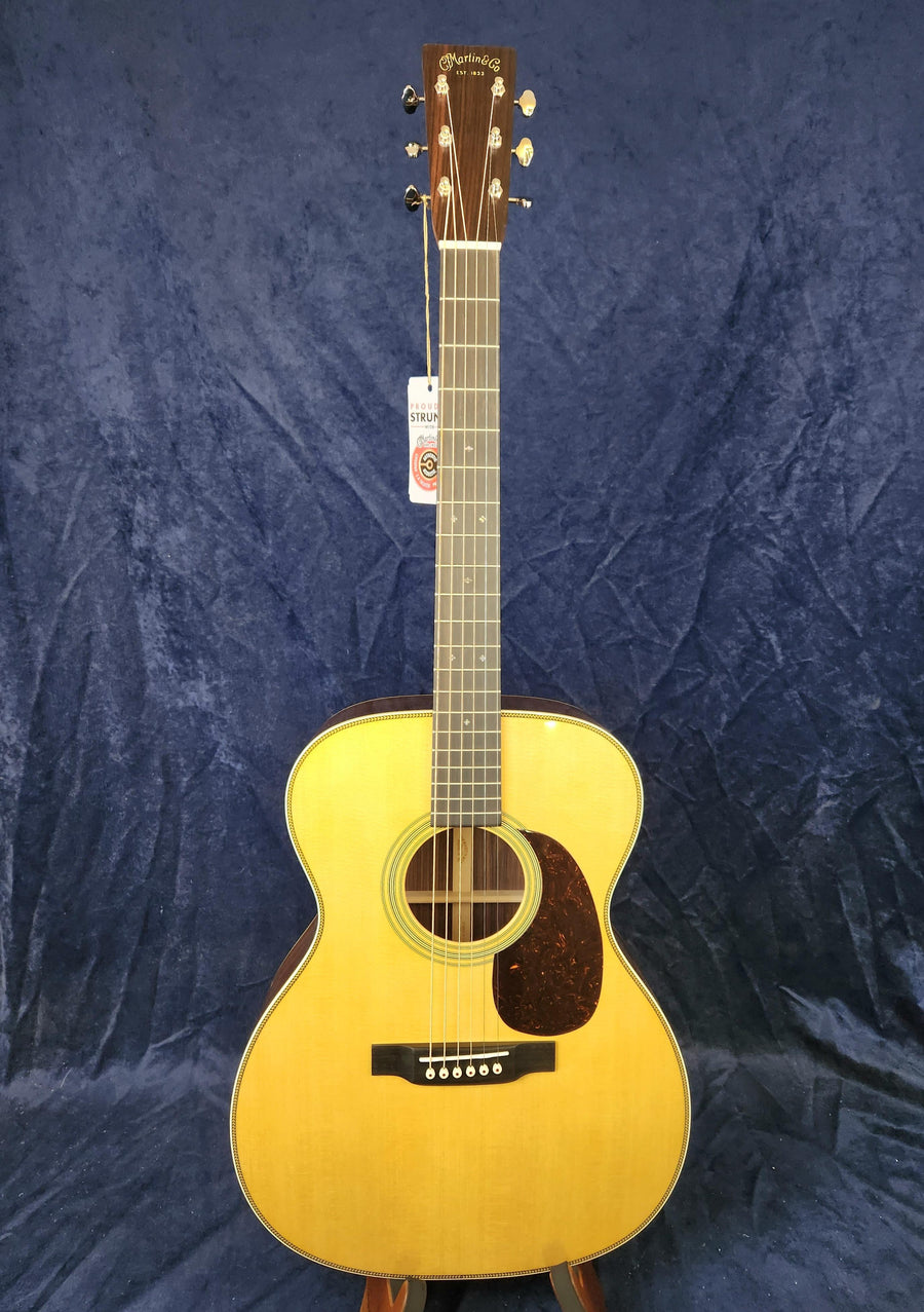 Light wood acoustic guitar with a dark pickguard, six strings, and a tag hanging from the headstock, set against a dark blue textured background.
