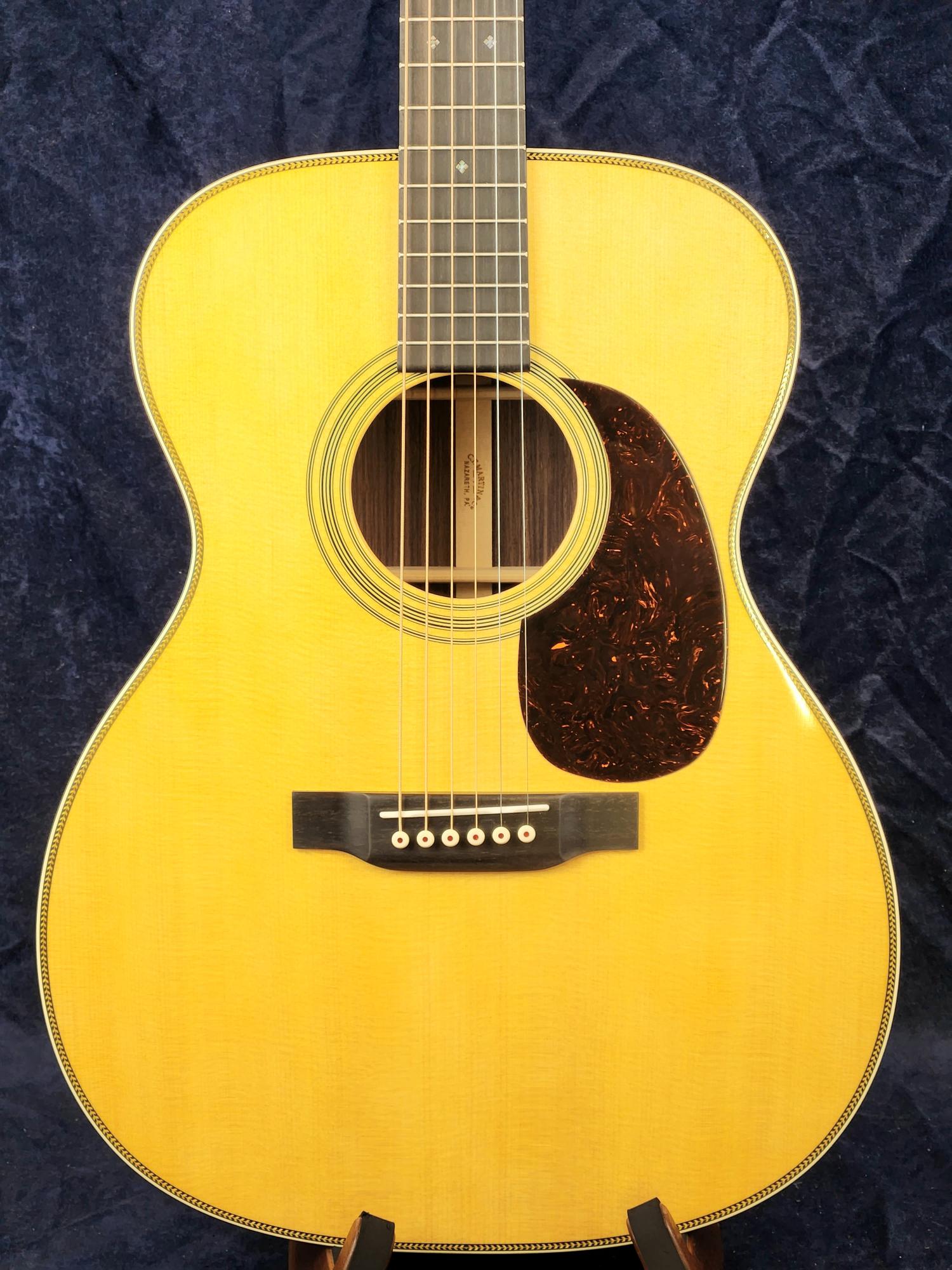 Light wood acoustic guitar with a dark tortoiseshell-style pickguard, decorative soundhole ring, and black bridge with six white bridge pins.