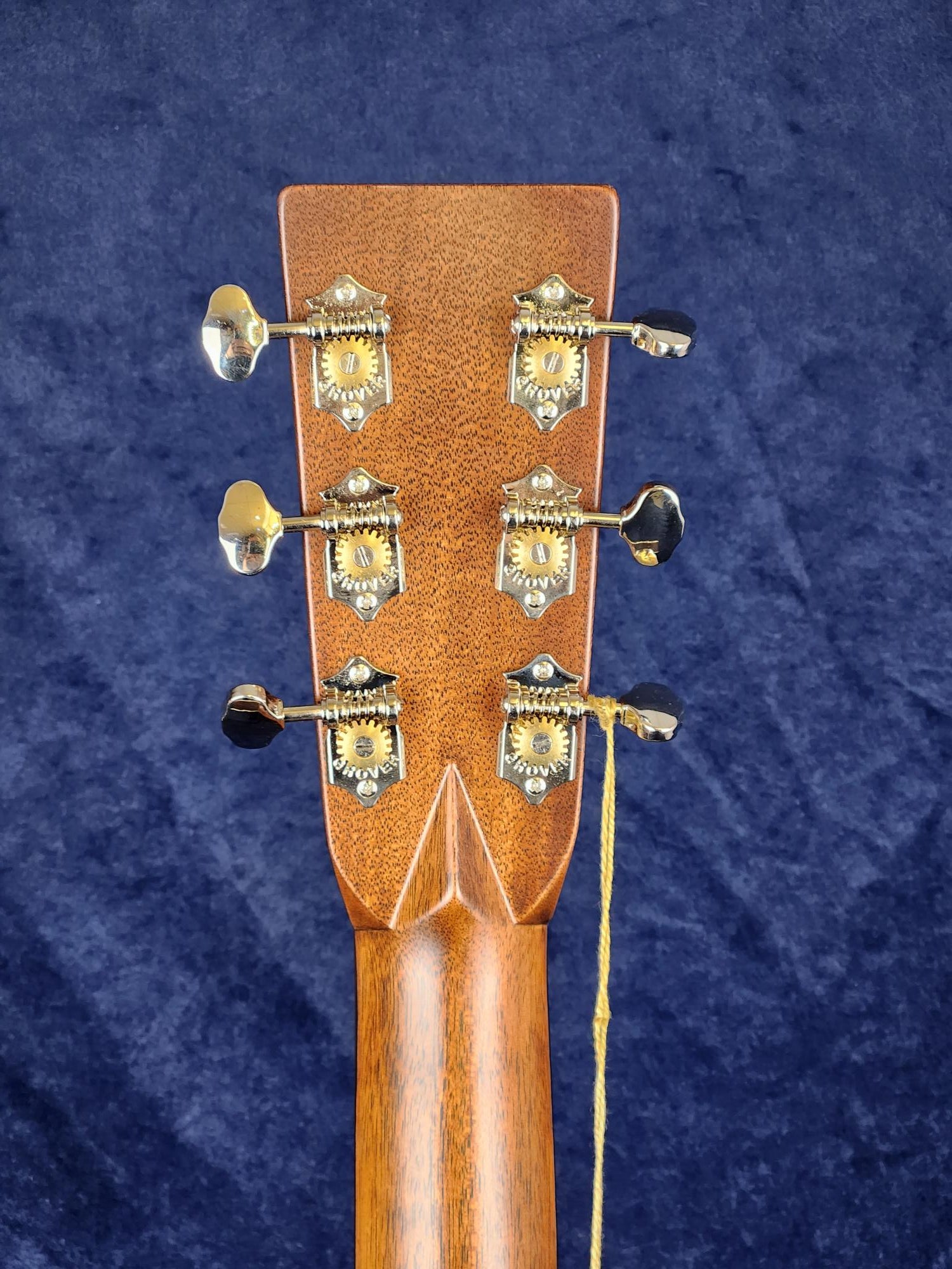 Six metal tuning pegs with ornate plates on the wooden headstock of an acoustic guitar, string ends visible, background is dark blue fabric.