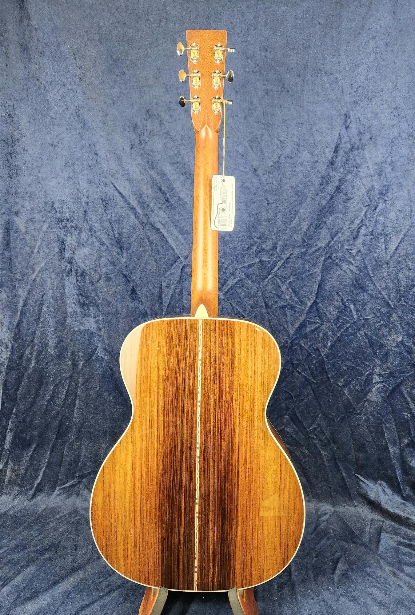 Acoustic guitar viewed from the back, showing glossy dark wood with a lighter center stripe and gold tuning pegs on the headstock.