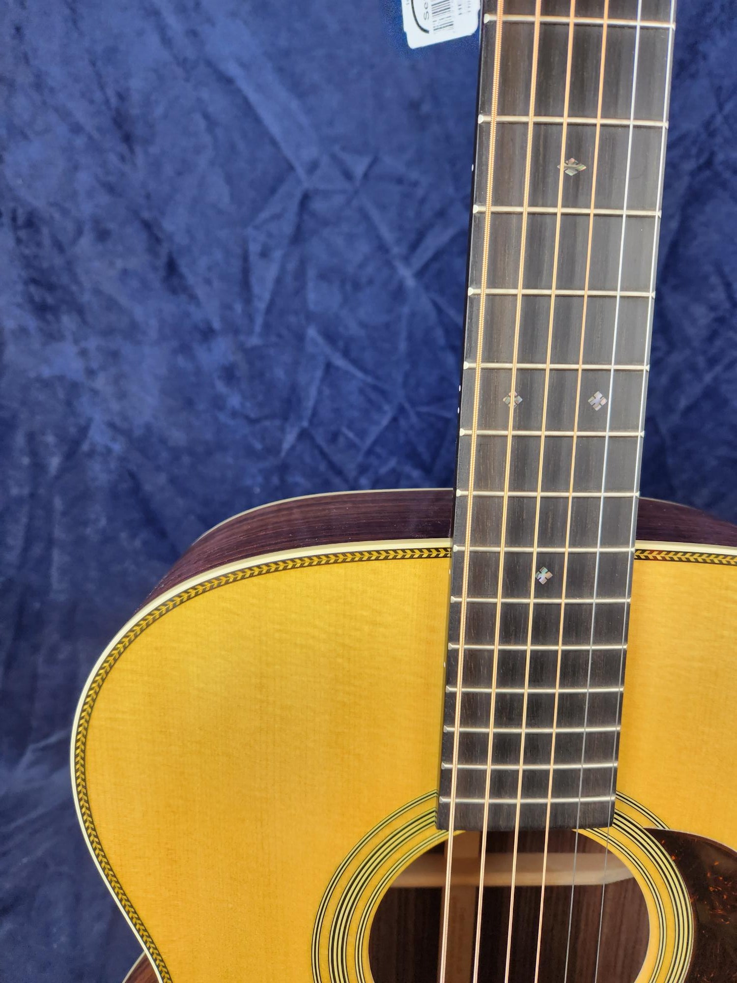 Light wood acoustic guitar with dark wood sides, diamond inlays on the fretboard, and decorative trim outlining the body against a blue textured background.