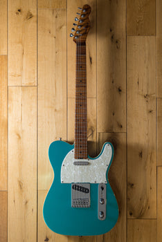 A teal electric guitar with a white pearloid pickguard, six tuning pegs, chrome hardware, and a maple neck, lying on a light wooden floor.