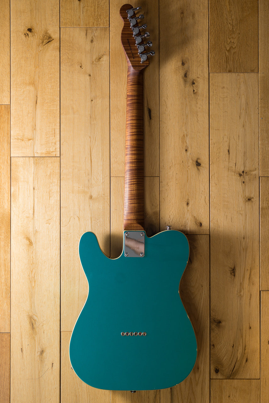 A teal electric guitar lying facedown on a light wooden floor, showing a striped wood neck and six tuning pegs.