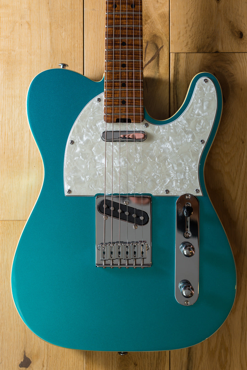 A teal electric guitar with a white pearloid pickguard, chrome hardware, and a brown wood neck, shown against a light wood background.