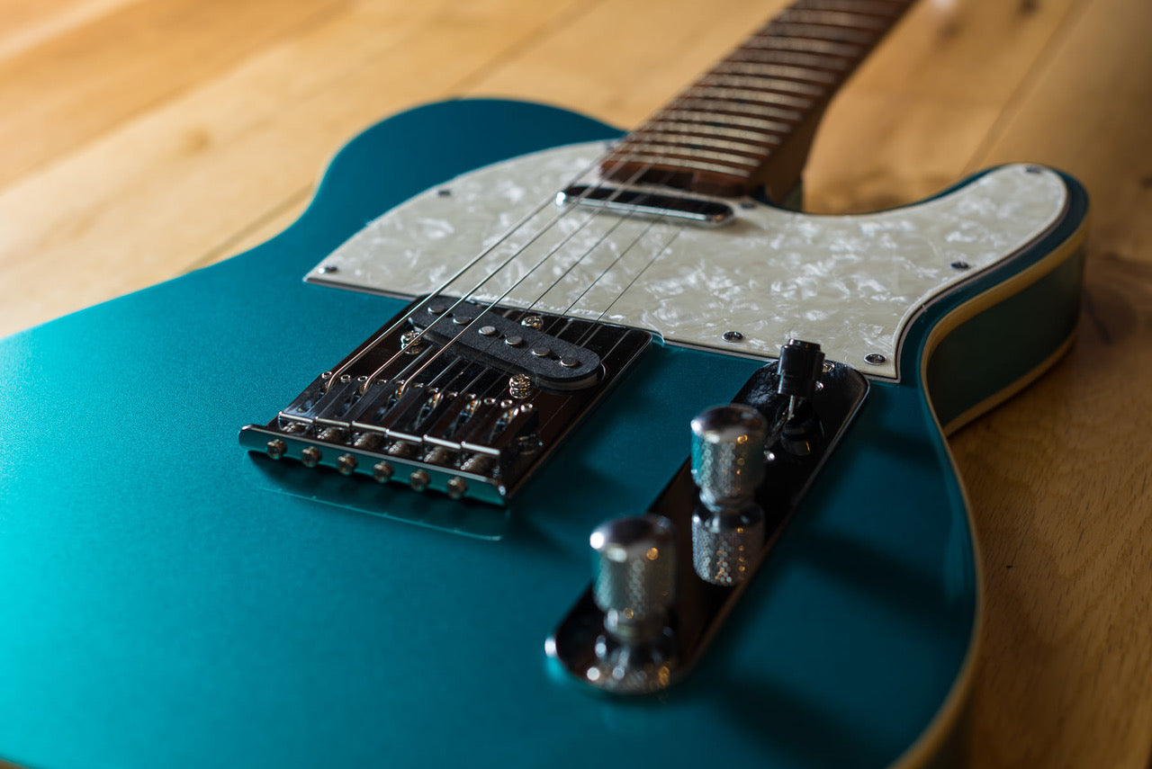 Teal electric guitar with a white pearloid pickguard, chrome bridge, two control knobs, and six strings, photographed on a wooden surface.