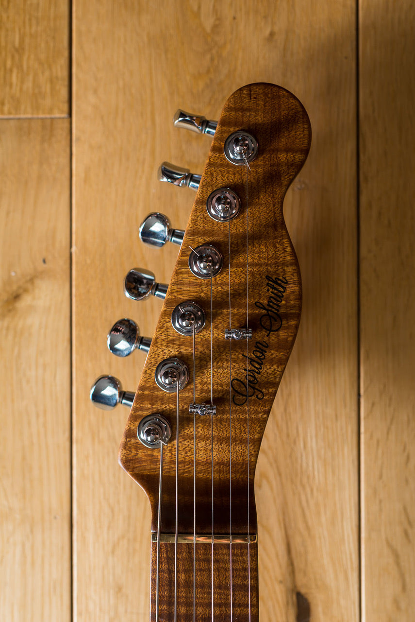 Electric guitar headstock in glossy brown wood with six silver tuning pegs and the words "Gordon Smith" written in black script.