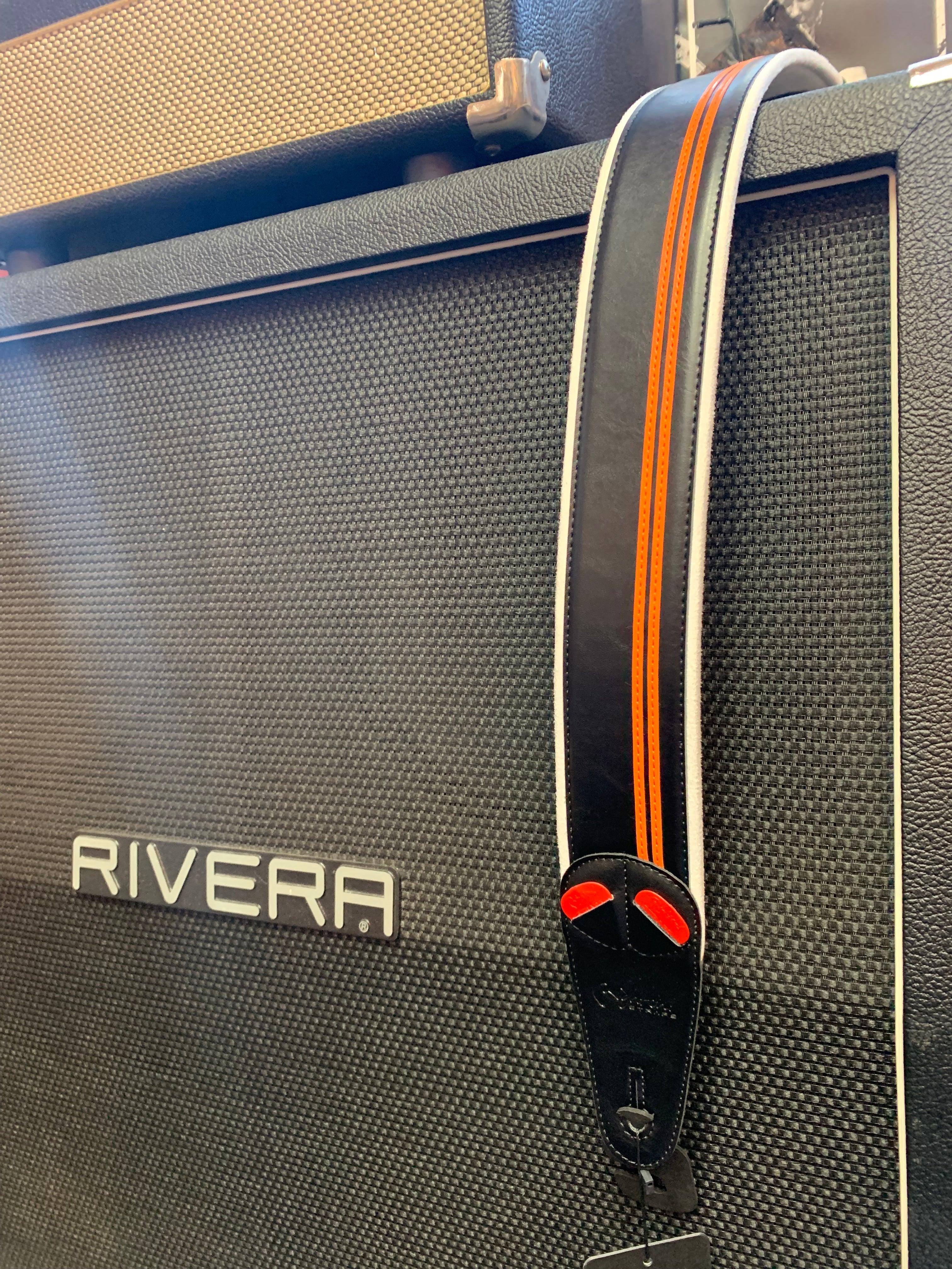 A black and orange guitar strap with red accents hangs over a large Rivera amplifier cabinet with a textured front panel.