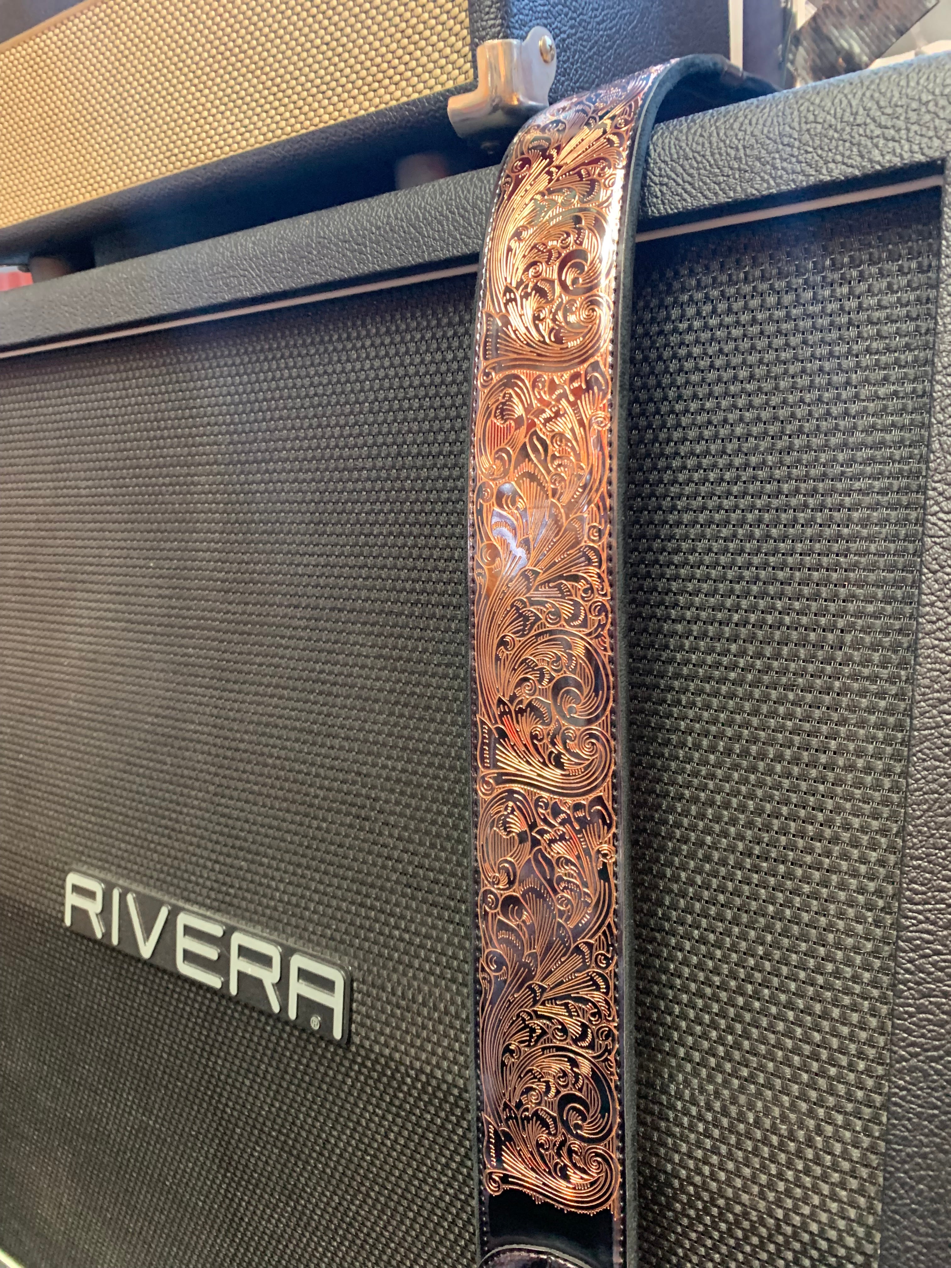 A leather guitar strap with intricate floral tooling hangs over a black Rivera amplifier.