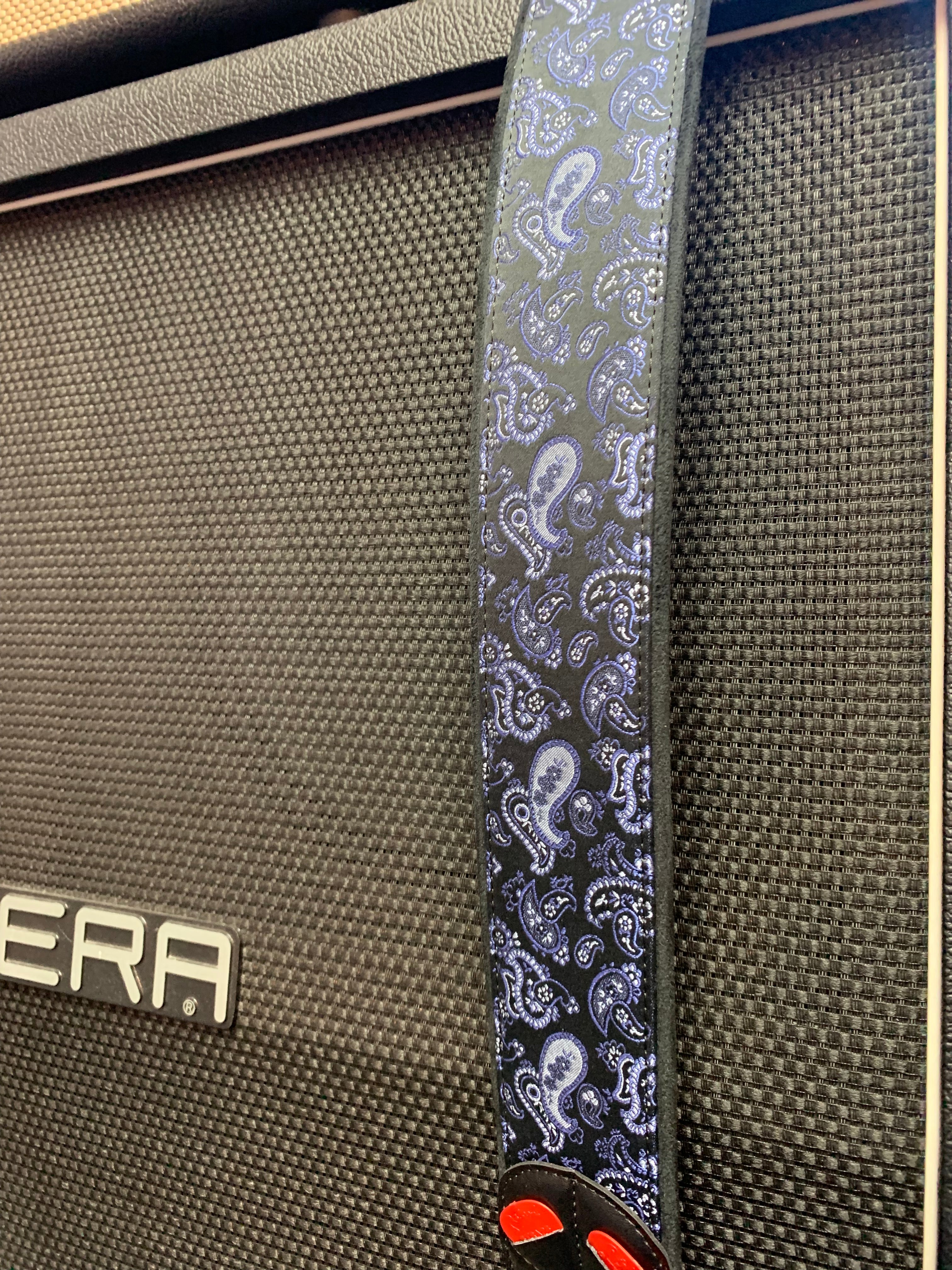 Black guitar amplifier with textured grille; a dark guitar strap featuring a blue paisley pattern hangs over the front.