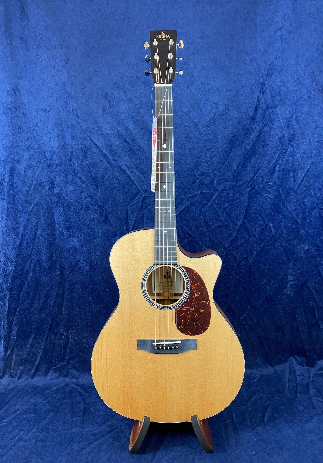 Sigma acoustic guitar with cutaway body, natural wood finish, dark pickguard, six strings, and decorative rosette, standing on a stand against blue cloth.