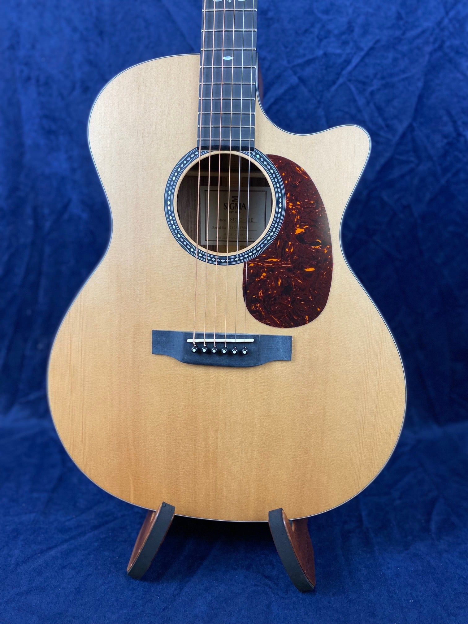 Light wood acoustic guitar with a single cutaway, tortoiseshell-style pickguard, and decorative rosette around the sound hole, displayed on a stand.